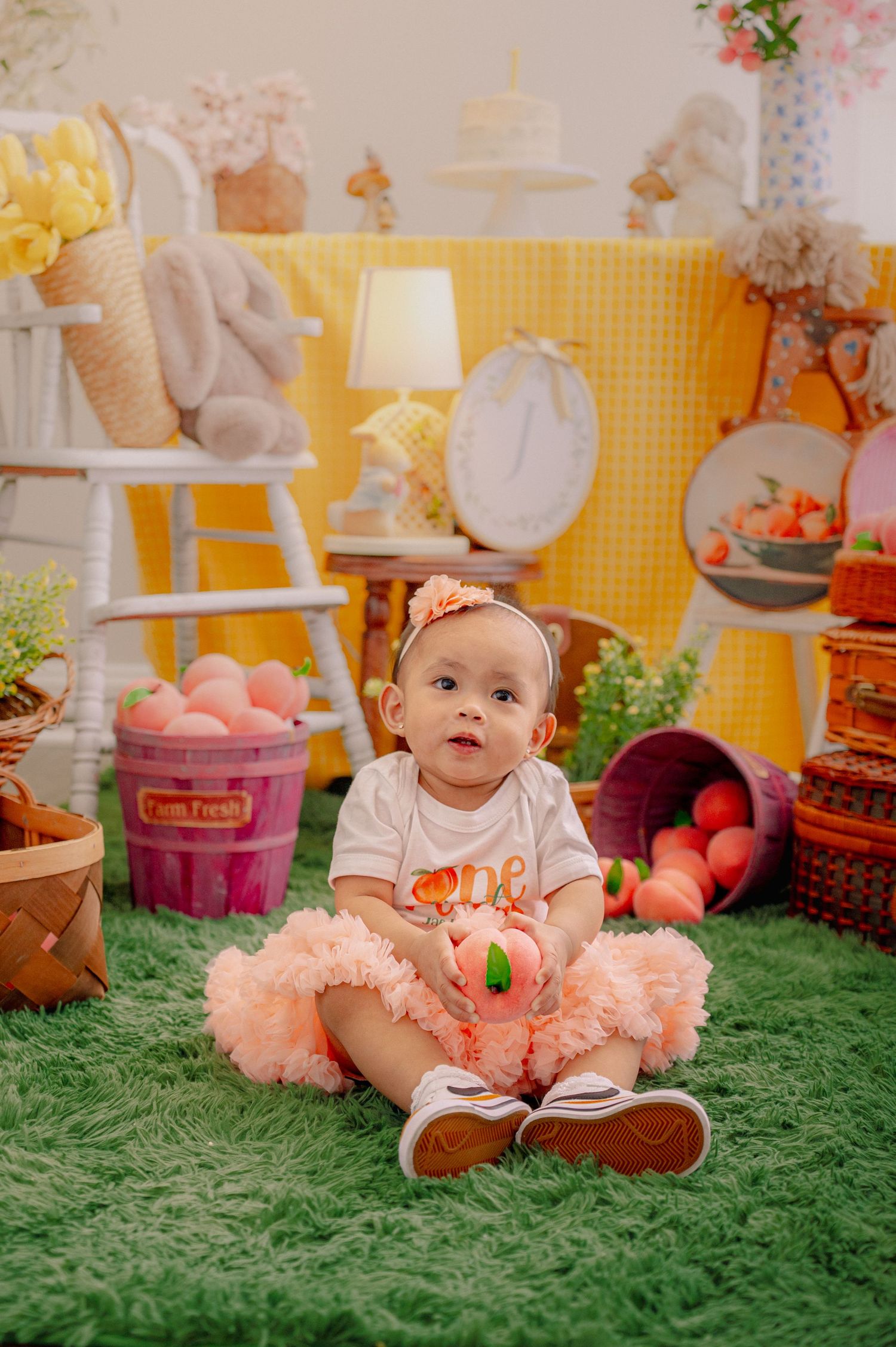 Baby in peach tutu dress sitting on green turf surrounded by party decorations.