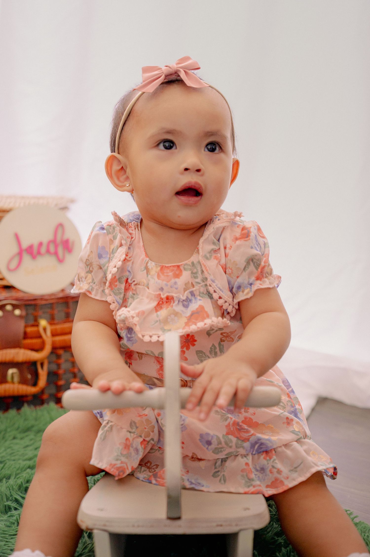 Baby in floral dress sitting on white chair during photo session.