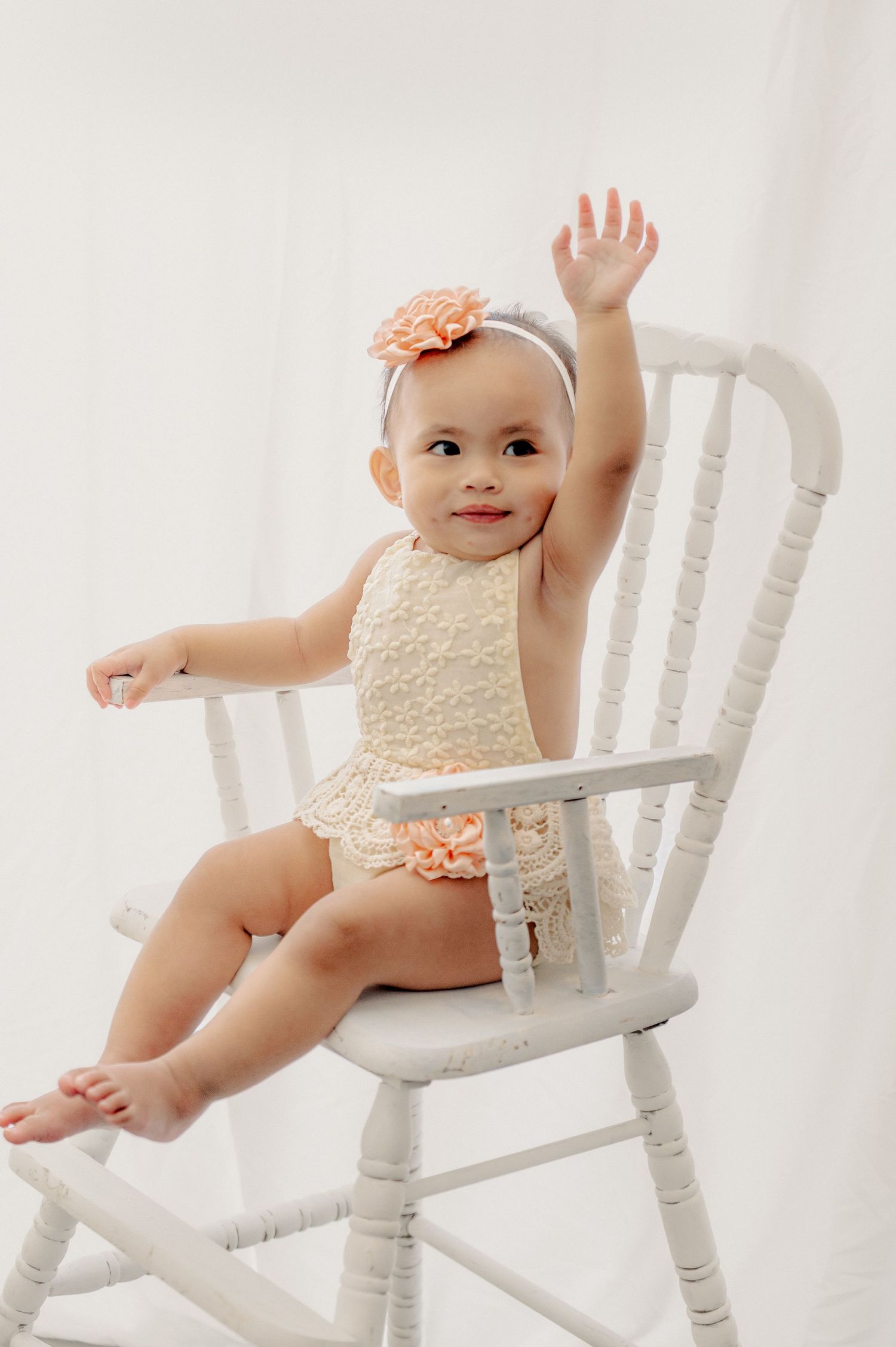 Baby in cream outfit posing on white wooden chair against white backdrop.