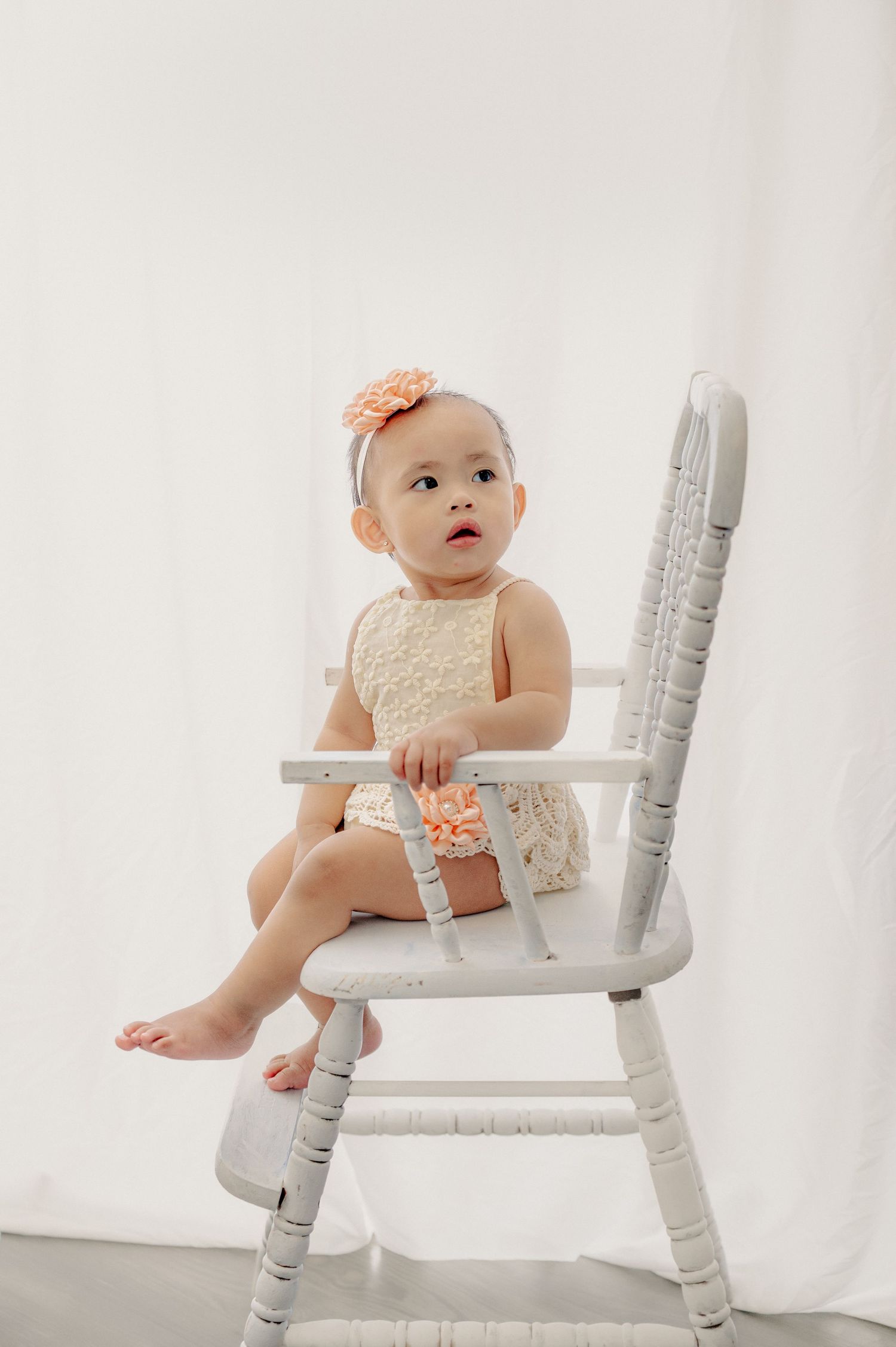 Baby in lace outfit sitting on vintage white wooden chair during photoshoot.