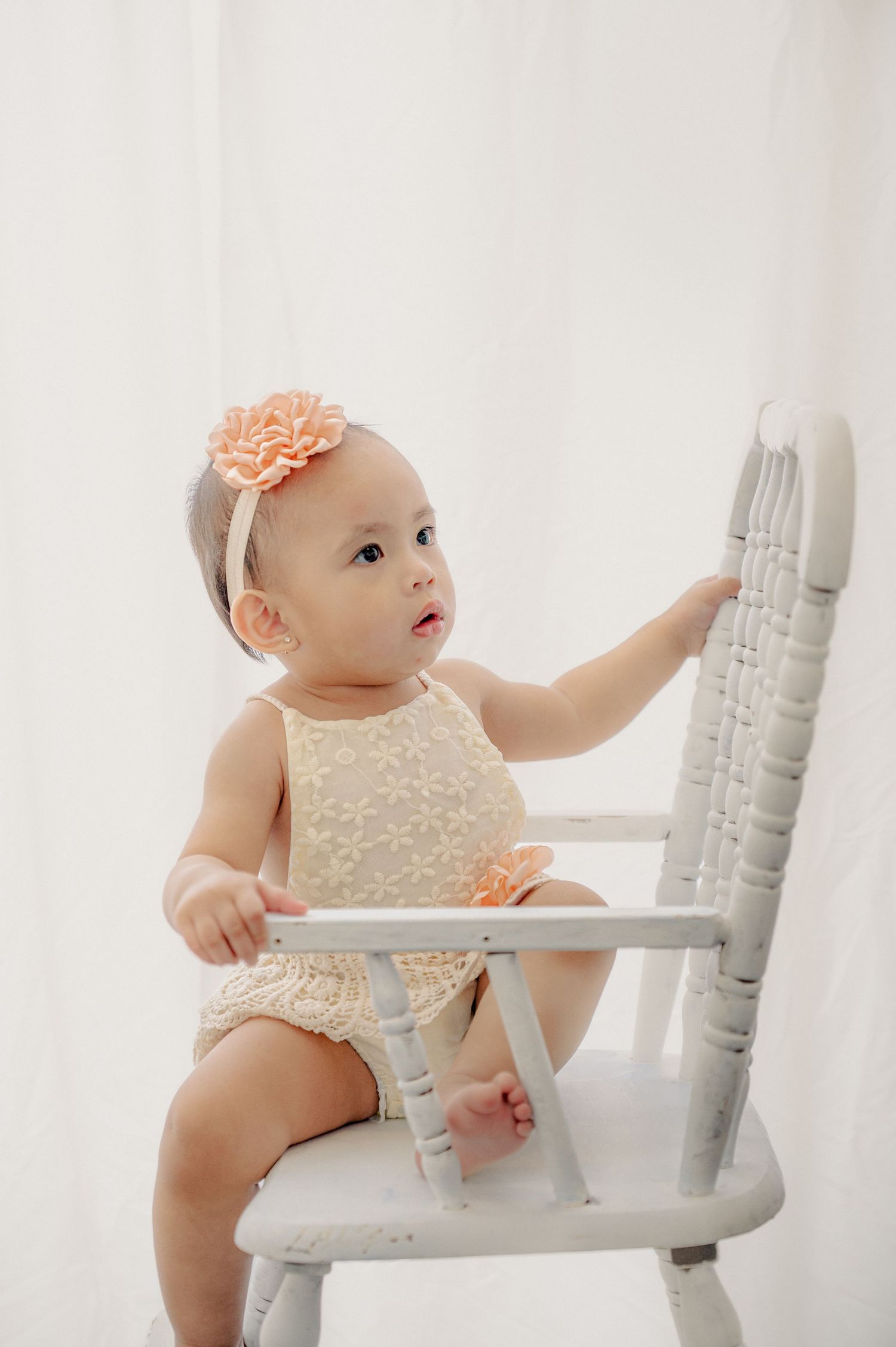 Baby in cream dress reaching up while seated on white wooden chair.