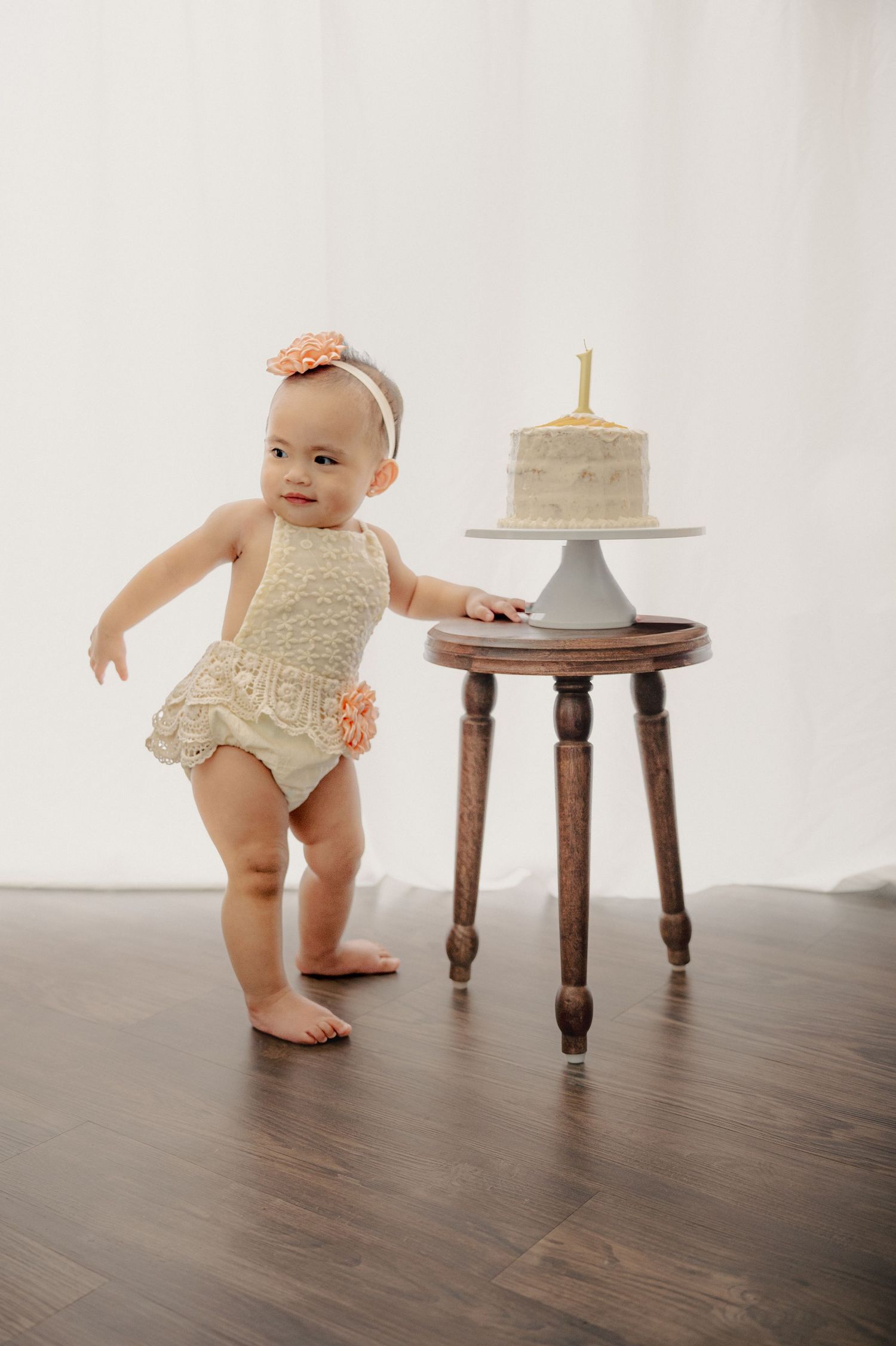Baby in a lace romper reaching towards a white cake displayed on a wooden stool against a light background.