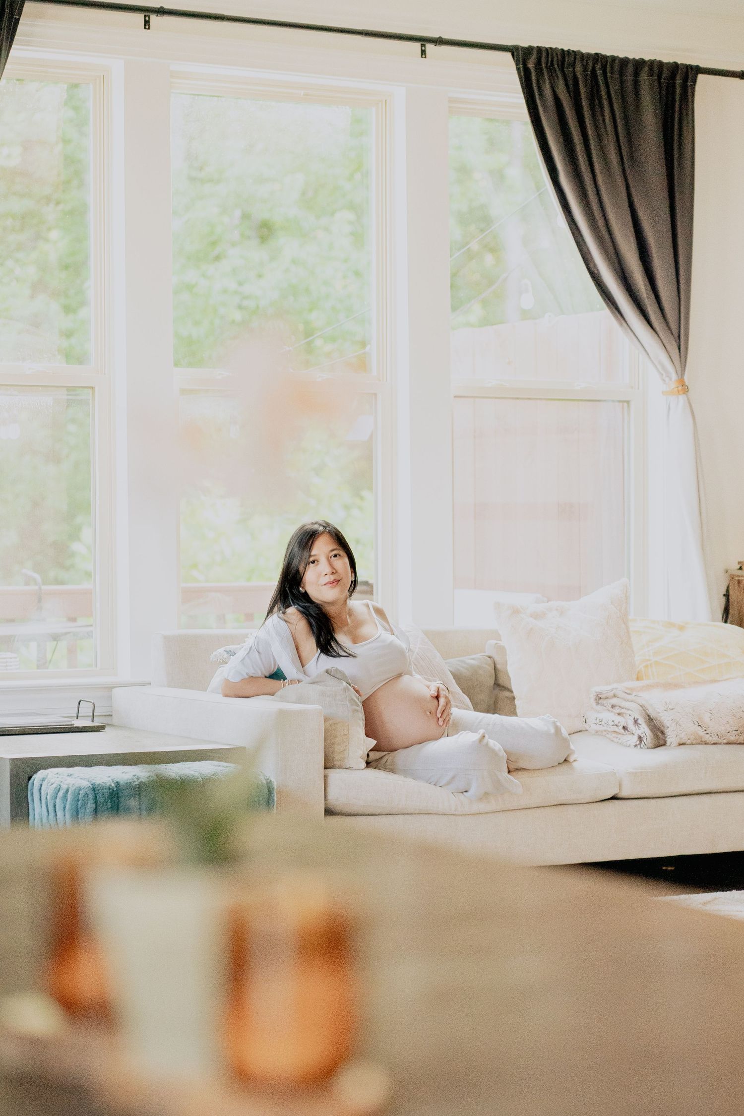 Pregnant woman relaxes on white couch near bright windows in serene living room setting.