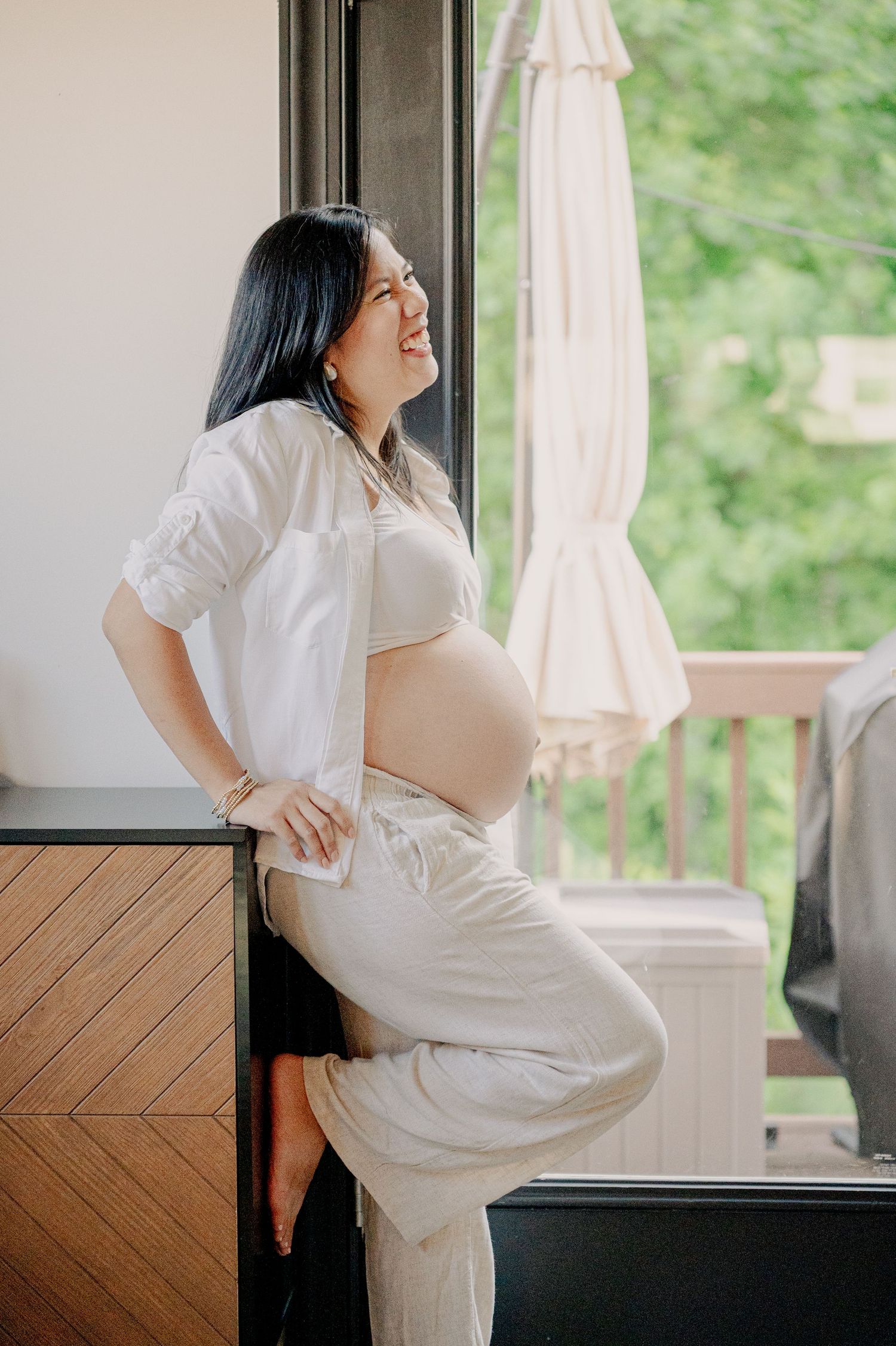 Maternity photoshoot in white dress by window with natural lighting.