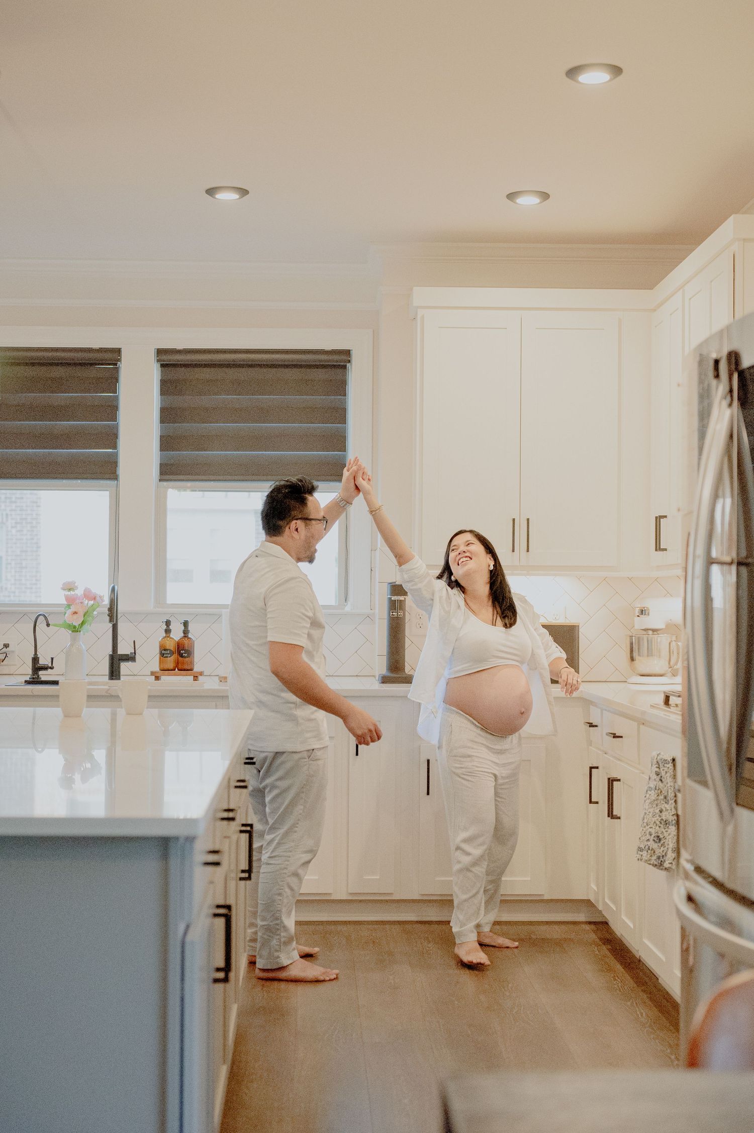 Couple shares romantic dance in white modern kitchen during maternity session.