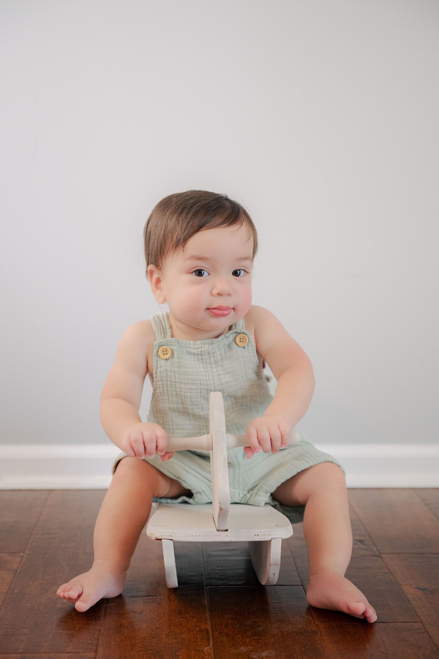 Baby in mint green overalls sits on a white rocking toy against a minimalist background.