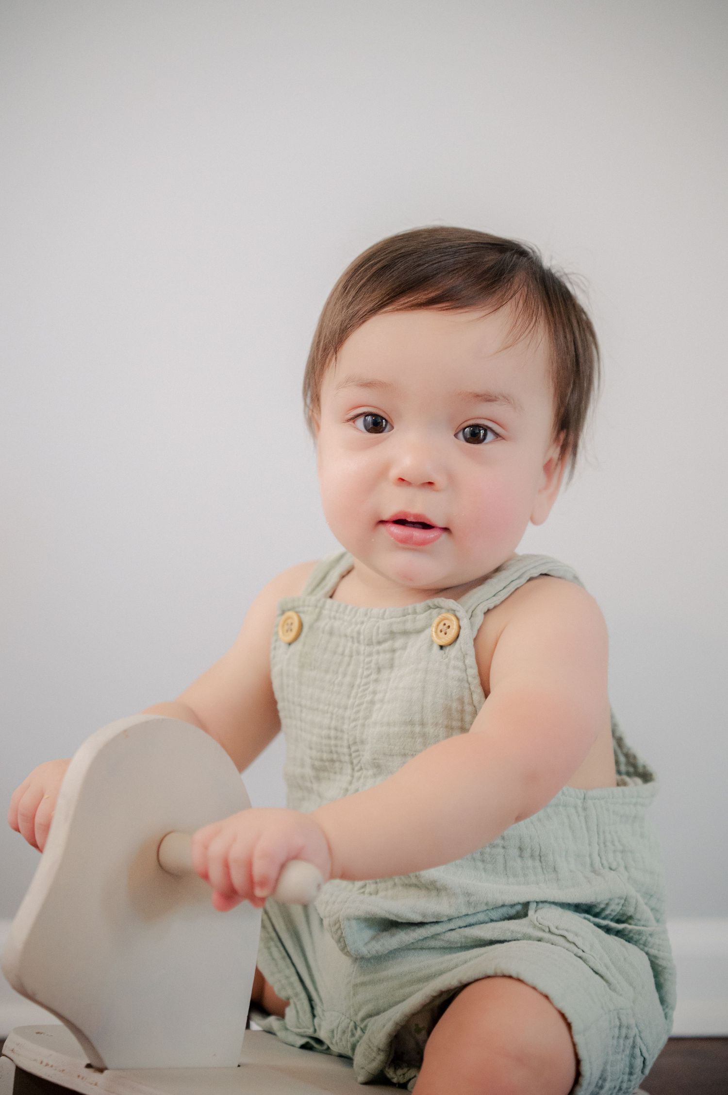 Baby in light green romper plays on a white rocking toy on hardwood floor.