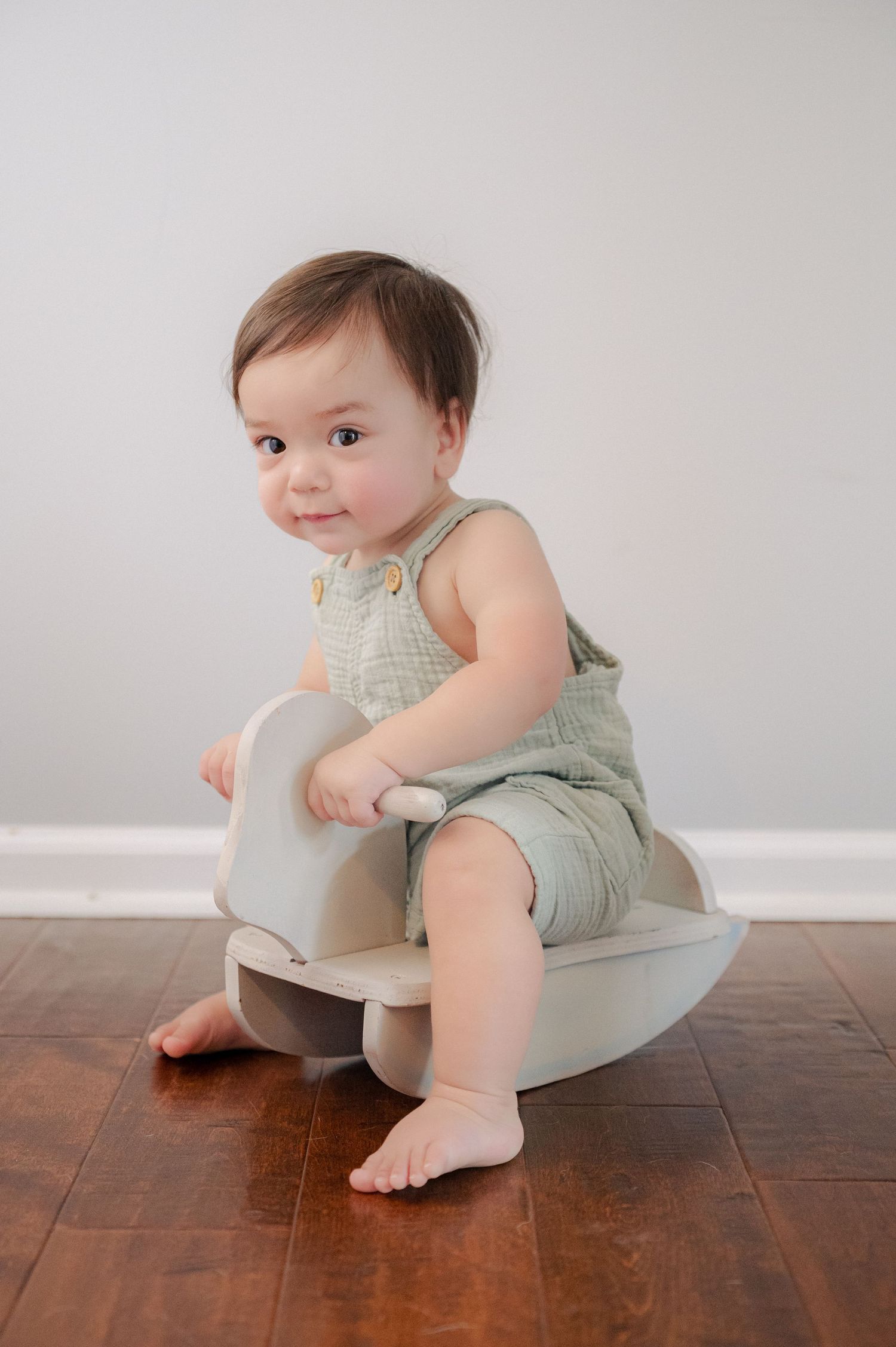 Small child wearing sage overalls enjoys playtime on a white rocker.