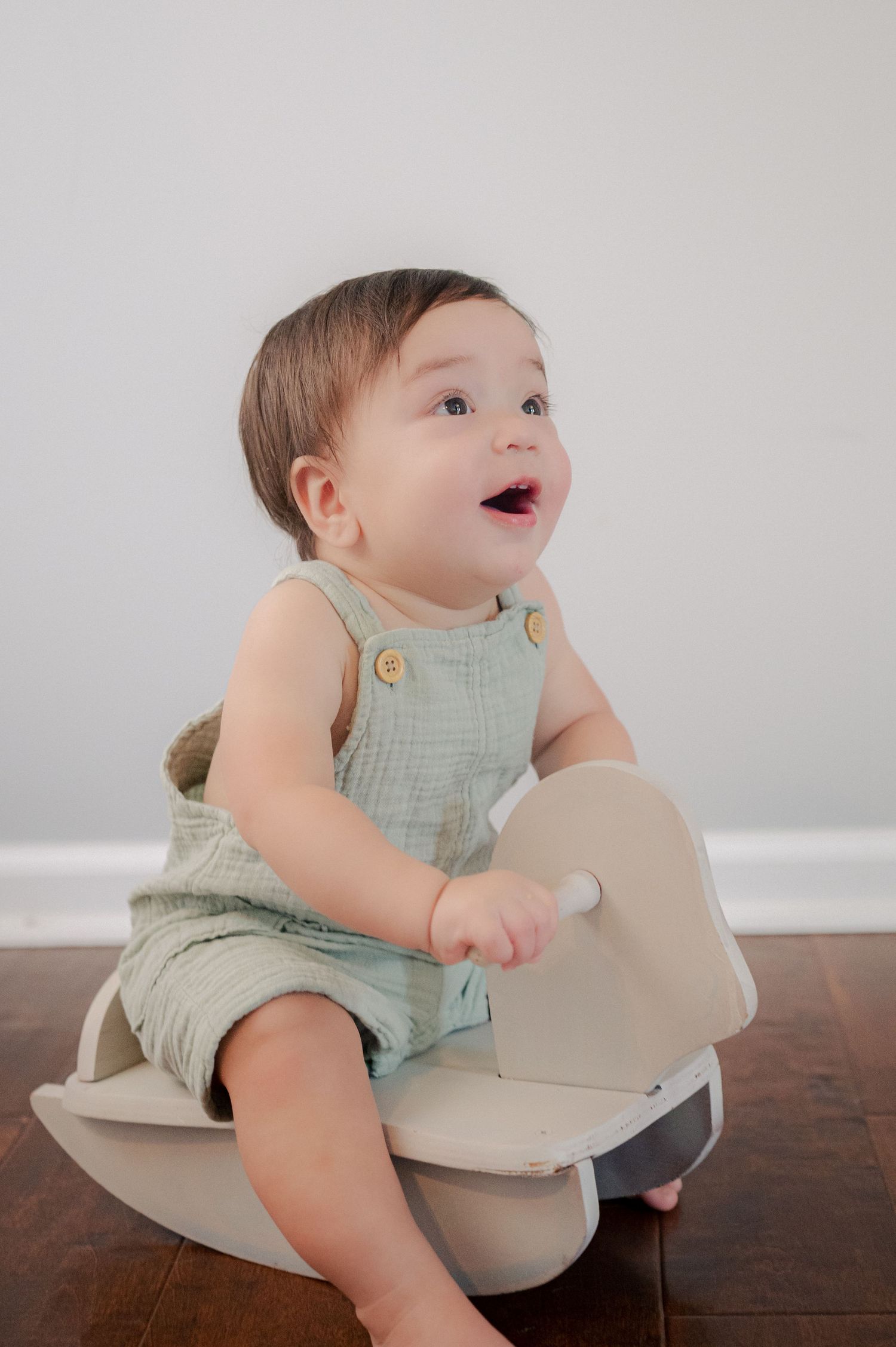 Baby in mint green romper sitting on beige cushion against white wall.