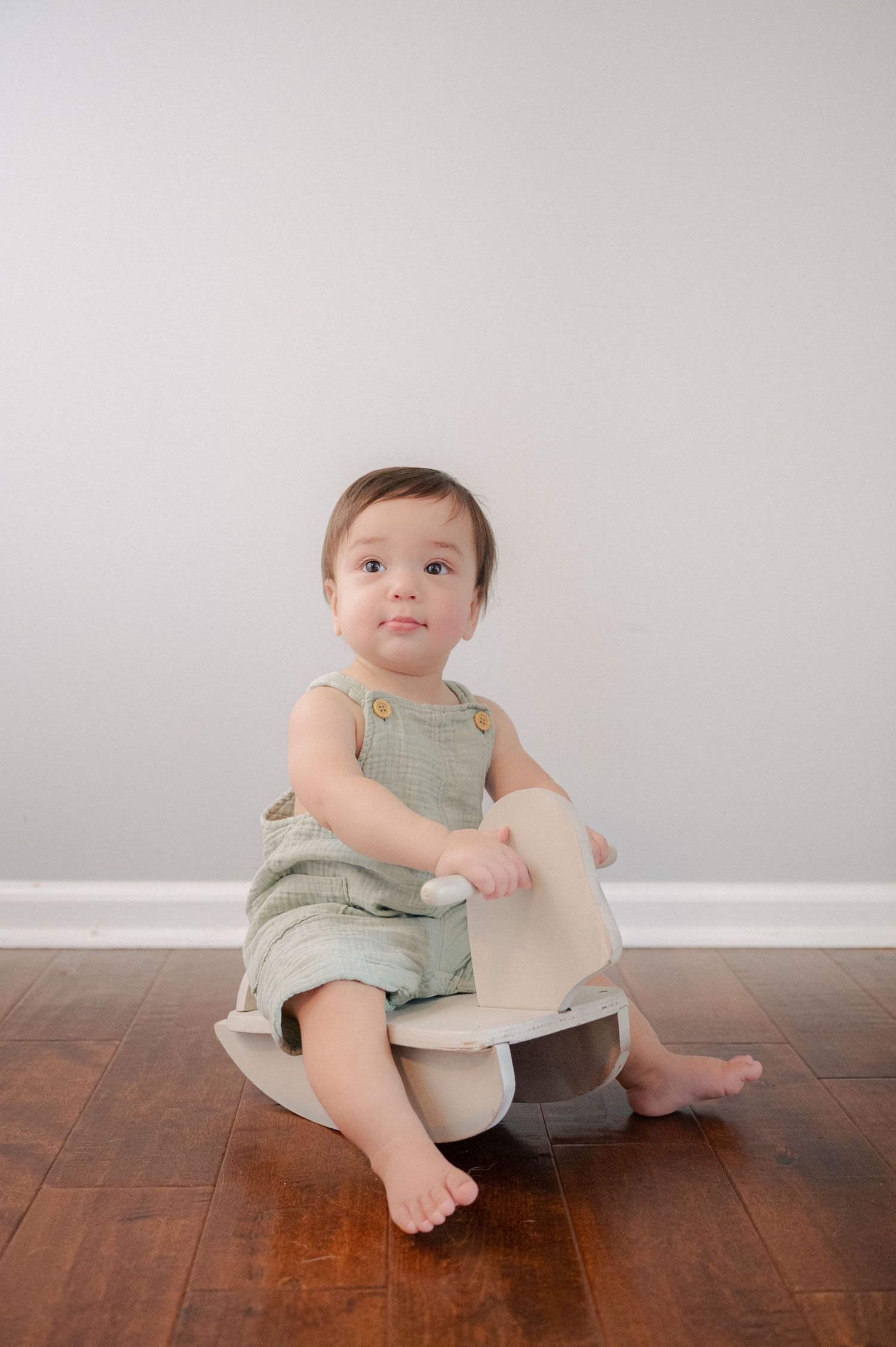 Baby in mint romper sits sideways on white rocking toy against neutral wall.