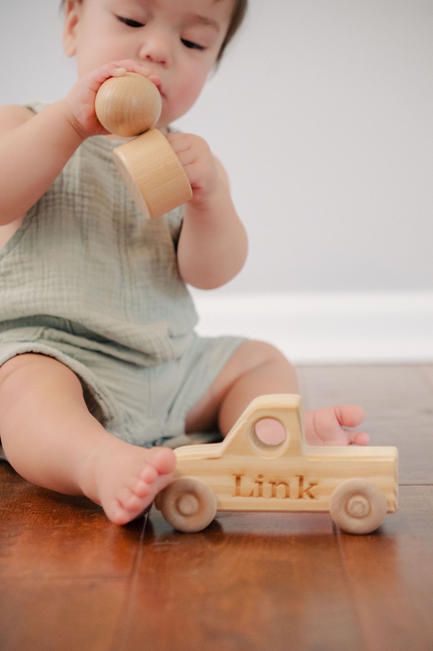 Baby plays with wooden toy truck labeled 'Link' while wearing mint green overalls.