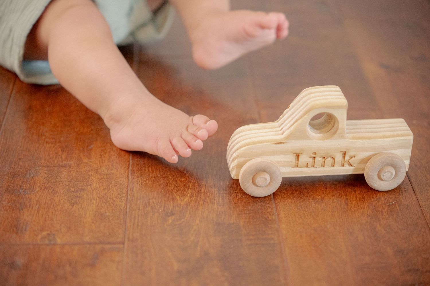Wooden toy truck with 'Link' engraving sits on hardwood floor next to small feet.