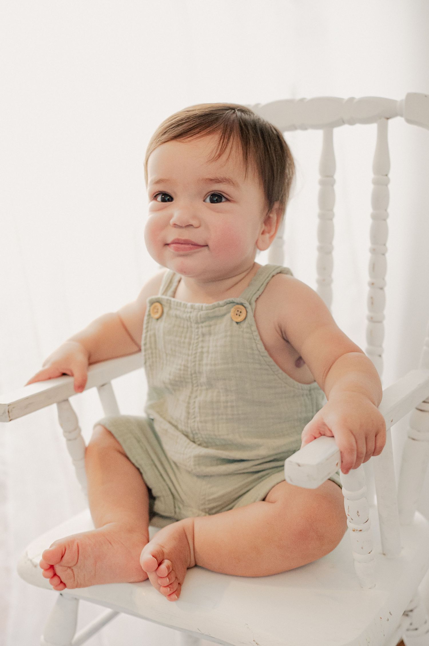 Baby in mint overalls sits on white vintage chair against light background.