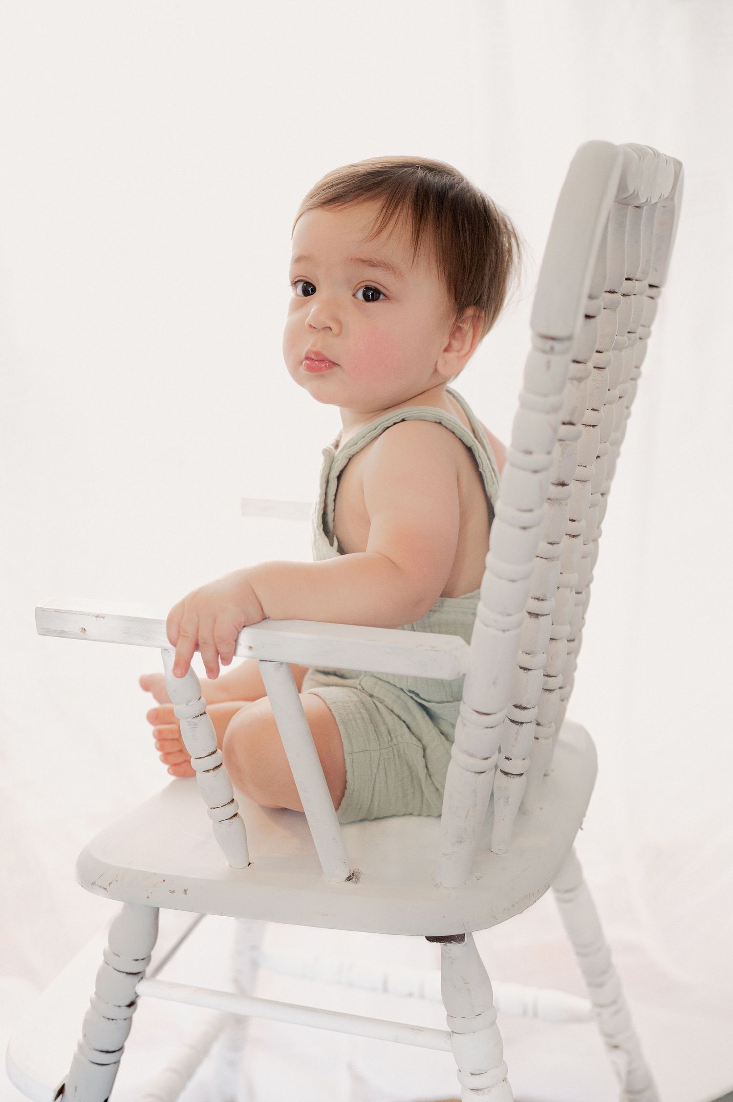Baby in sage romper sits on decorative white wooden chair during photo session.