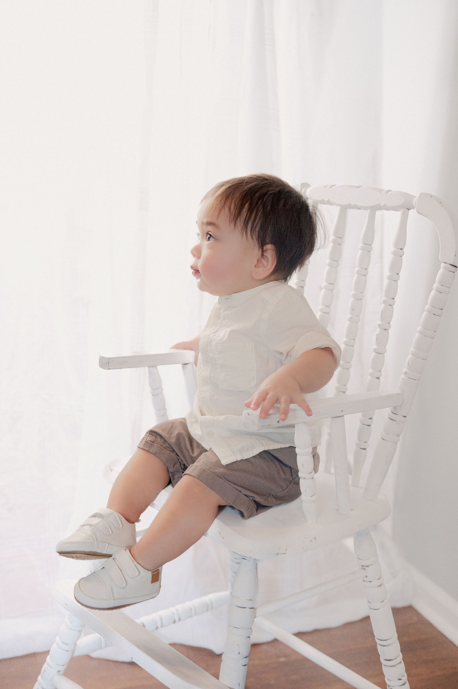 Small child in white shirt and shorts sits sideways on vintage white rocking chair.