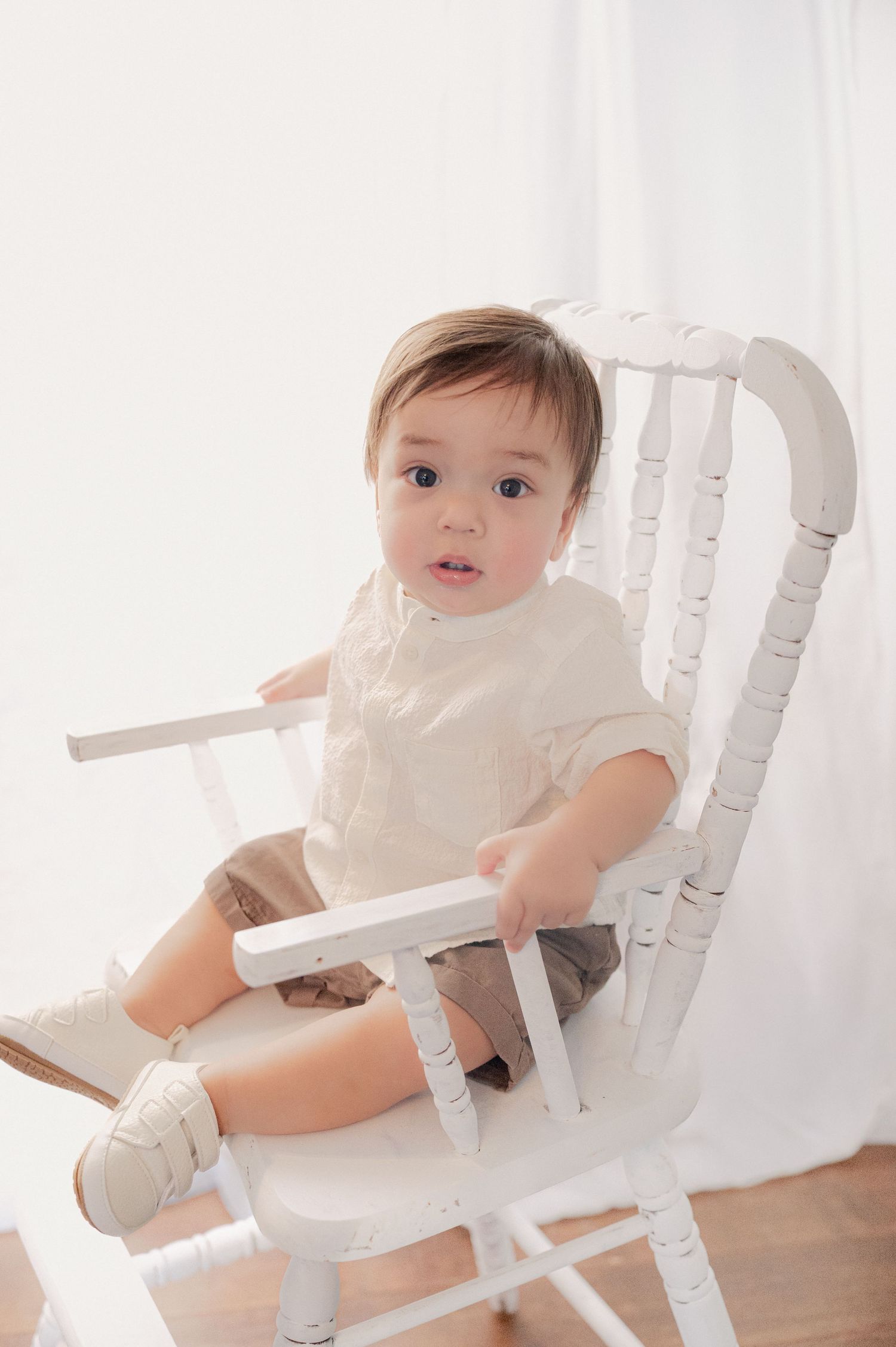 Baby sitting in white high chair wearing white outfit against light background.