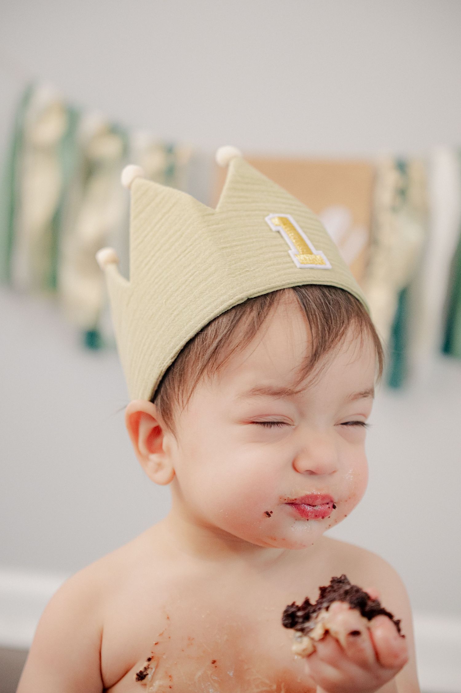 Smiling baby in yellow crown enjoying birthday cake with frosting on face.