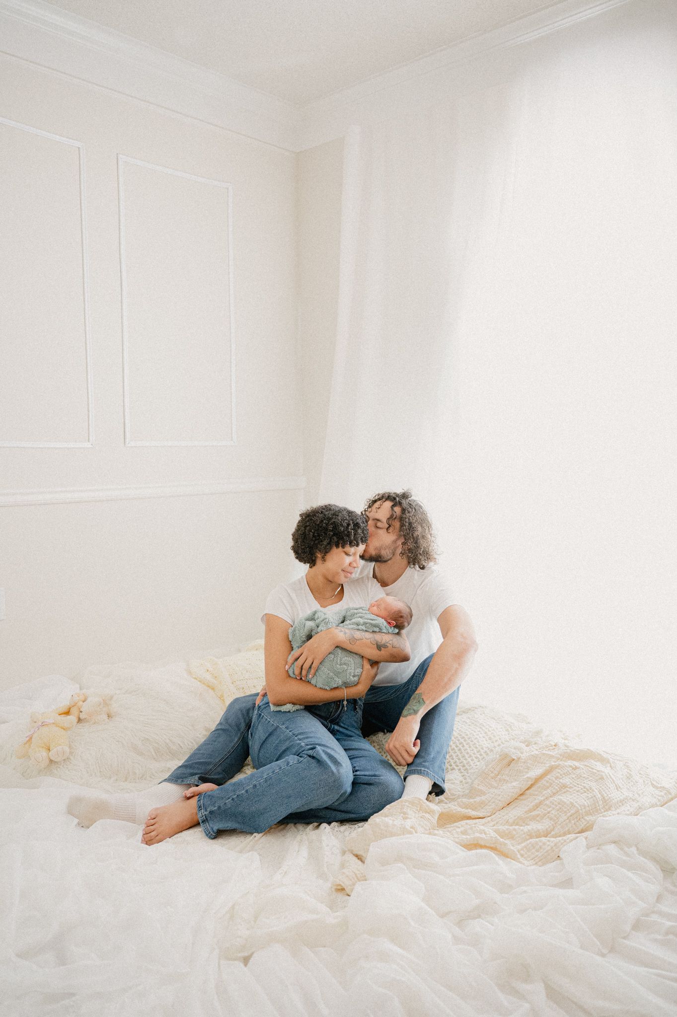 Couple sitting on white blanket cuddles with their newborn baby in minimalist setting.