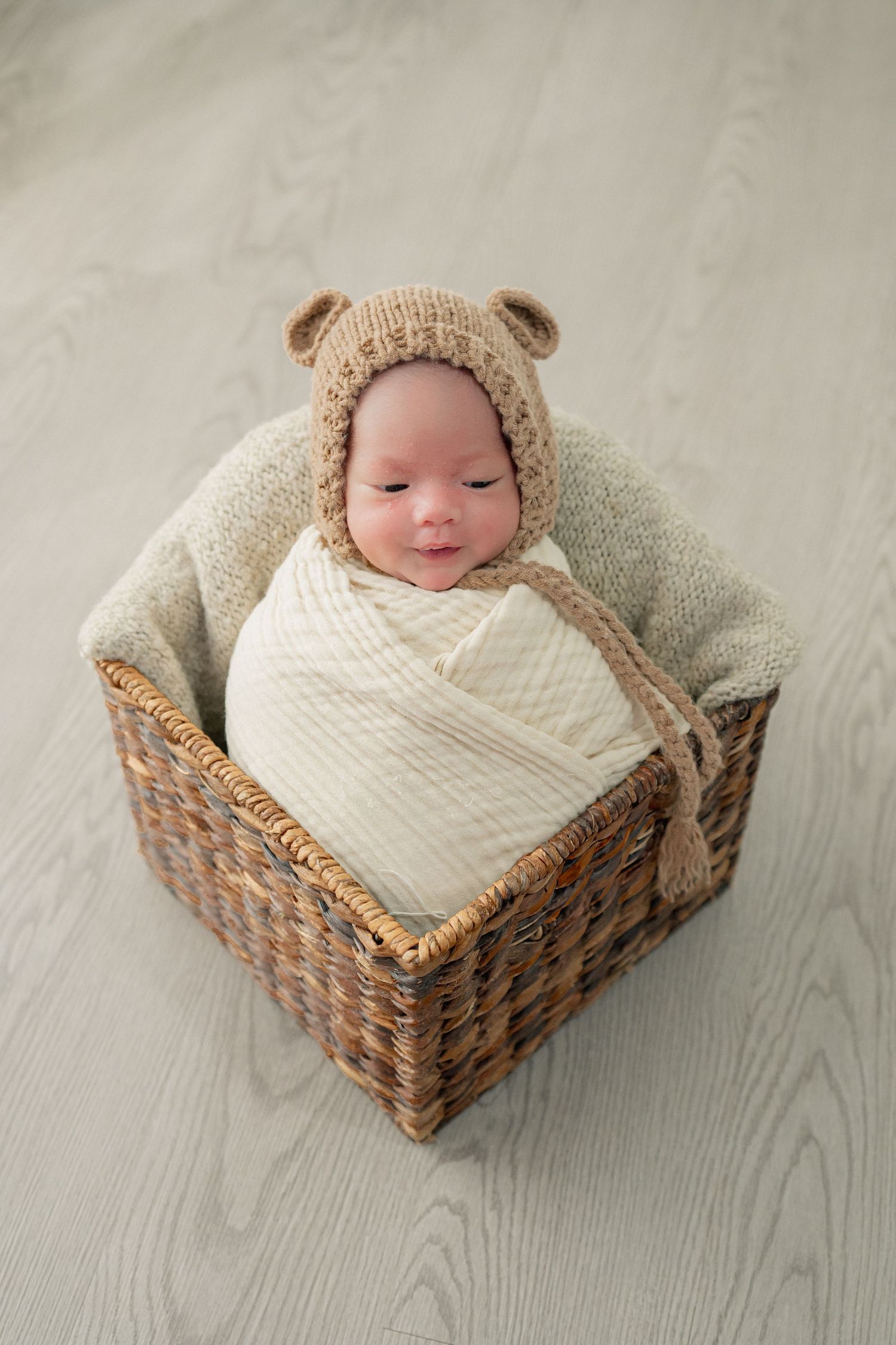 Sleeping newborn wrapped in white blanket lying in woven basket wearing knit bear hat.