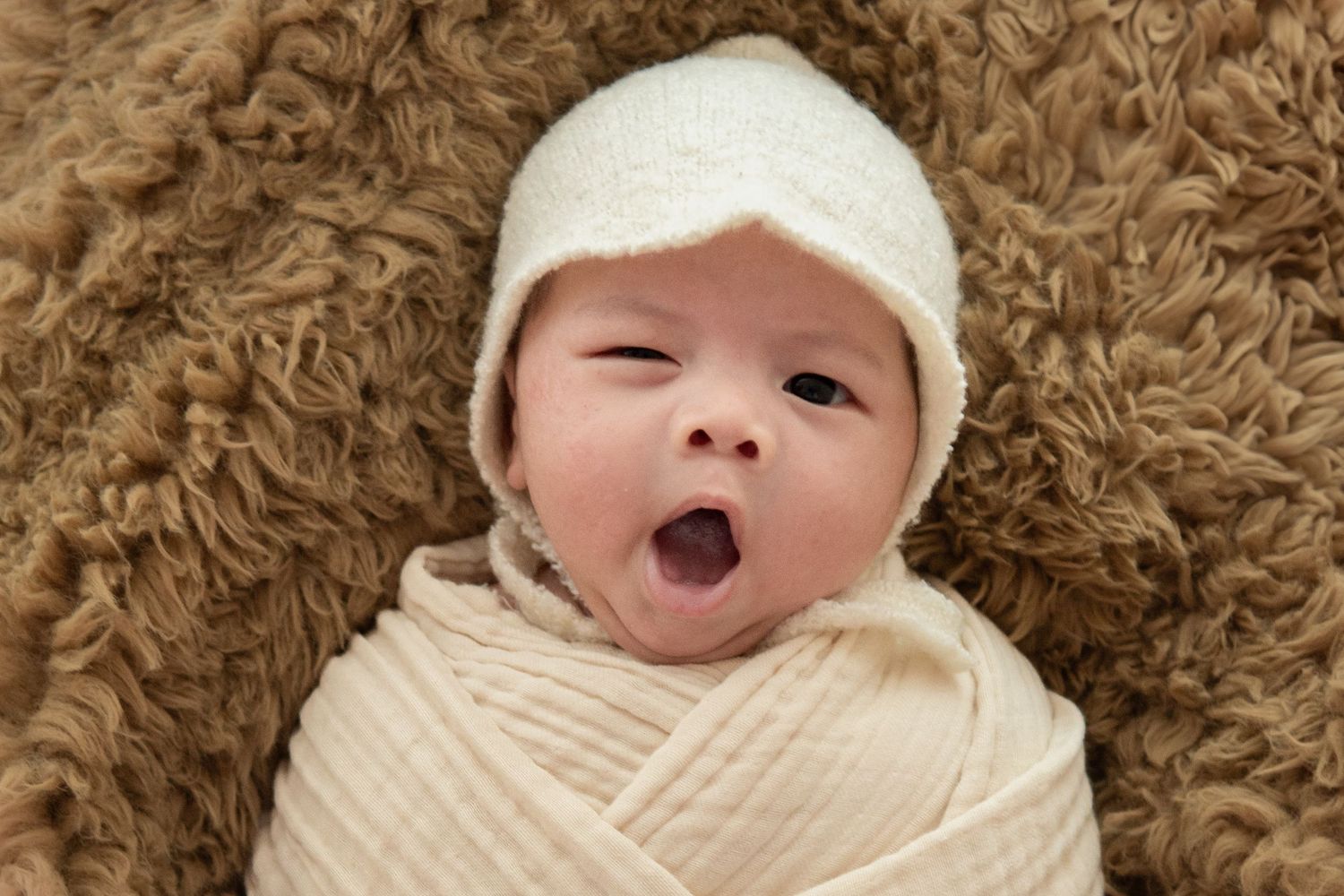 Alert newborn wearing white cap yawns while swaddled against brown textured background.