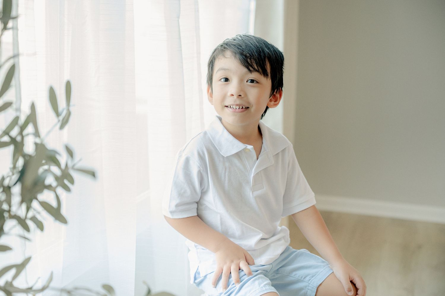 Child in white shirt sits by window in minimalist indoor setting with soft natural lighting.