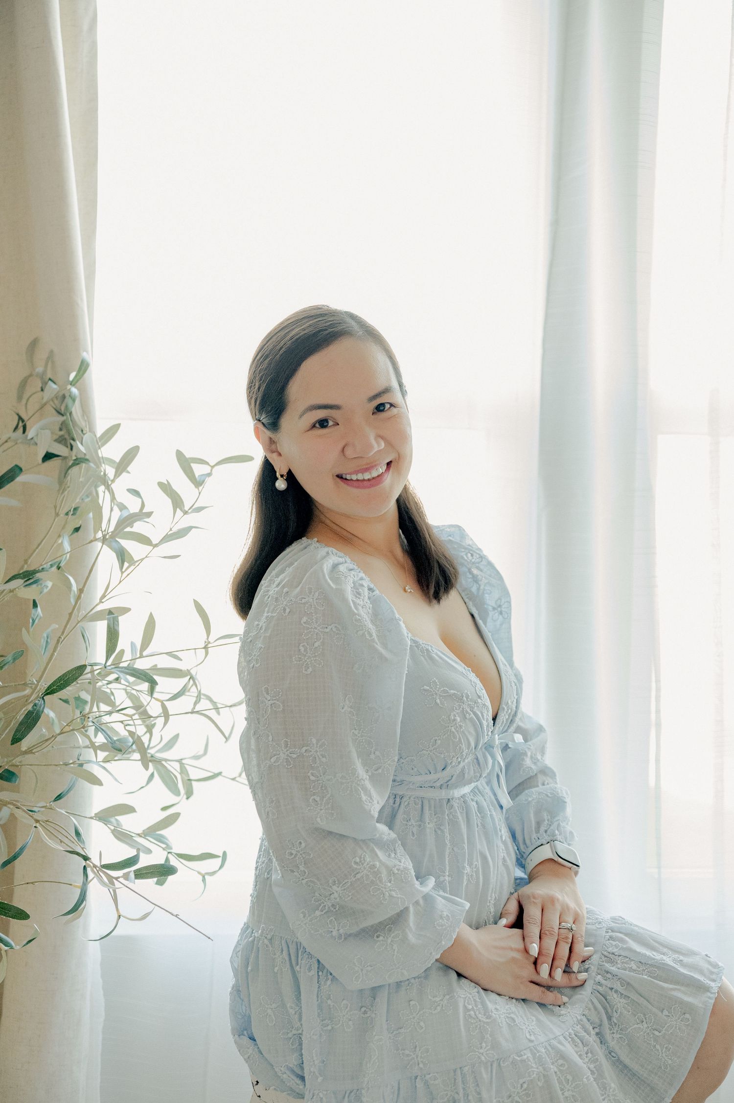 Woman in white dress sitting by window with soft natural light and greenery.
