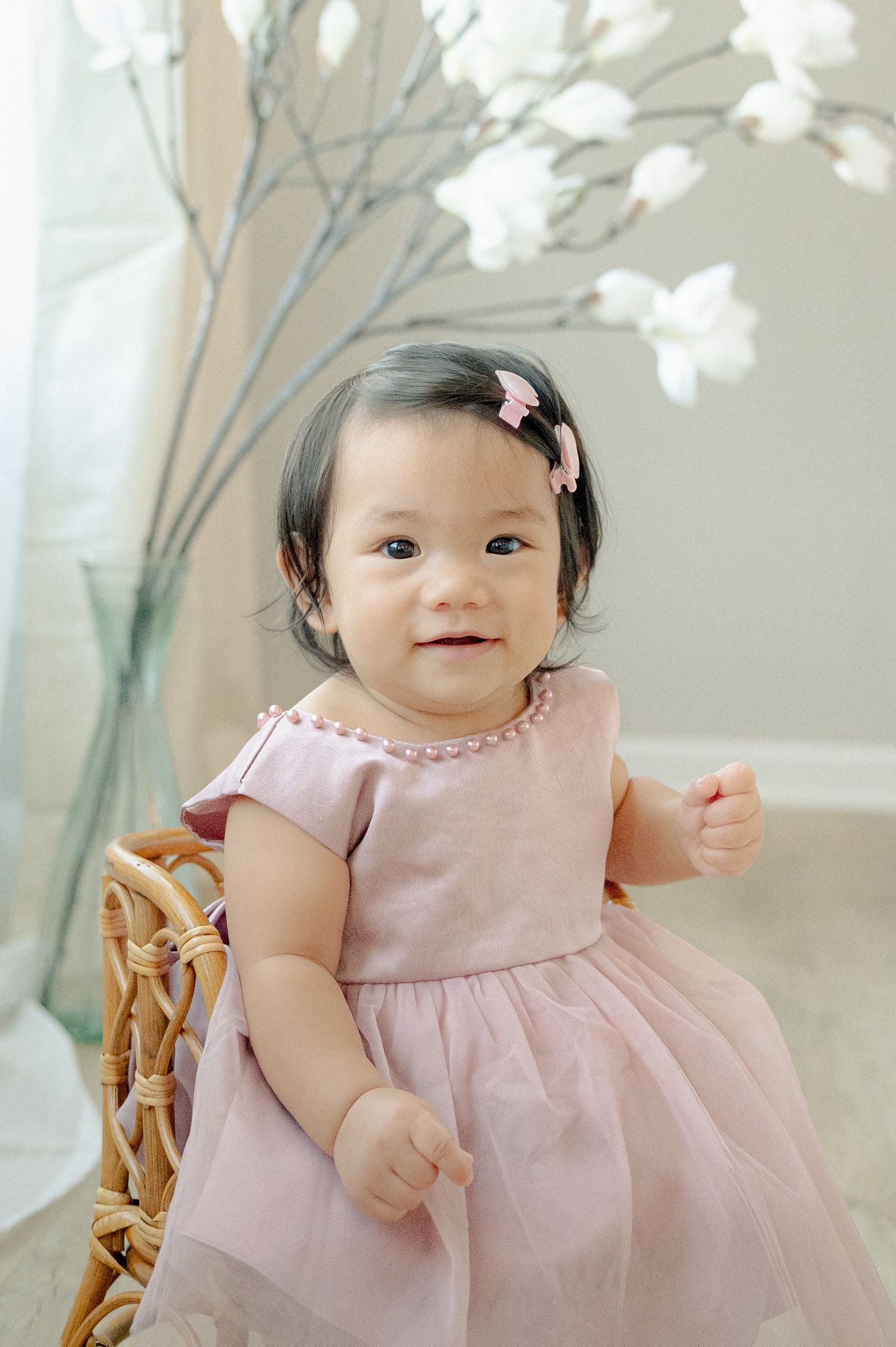 Baby in pink dress sitting on wooden chair with white floral decorations.