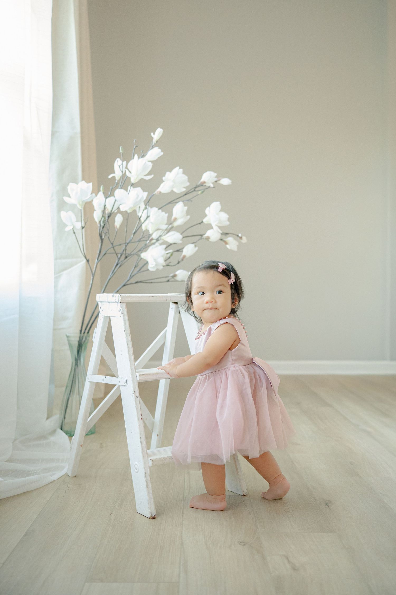 Toddler in pink tulle dress exploring white decorative elements on wooden floor.