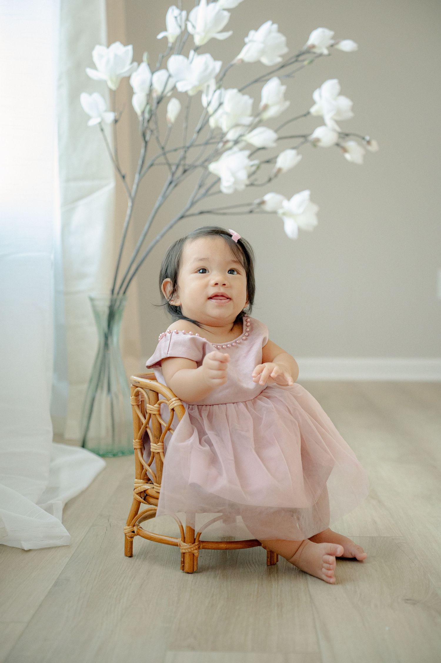 Baby in pink dress posing with white floral arrangements on light wooden floor.