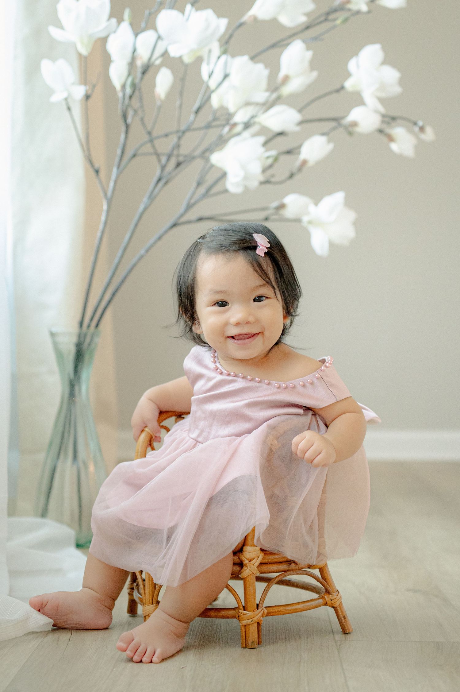 Happy baby in pink dress playing on small rattan chair with white flowers in background.