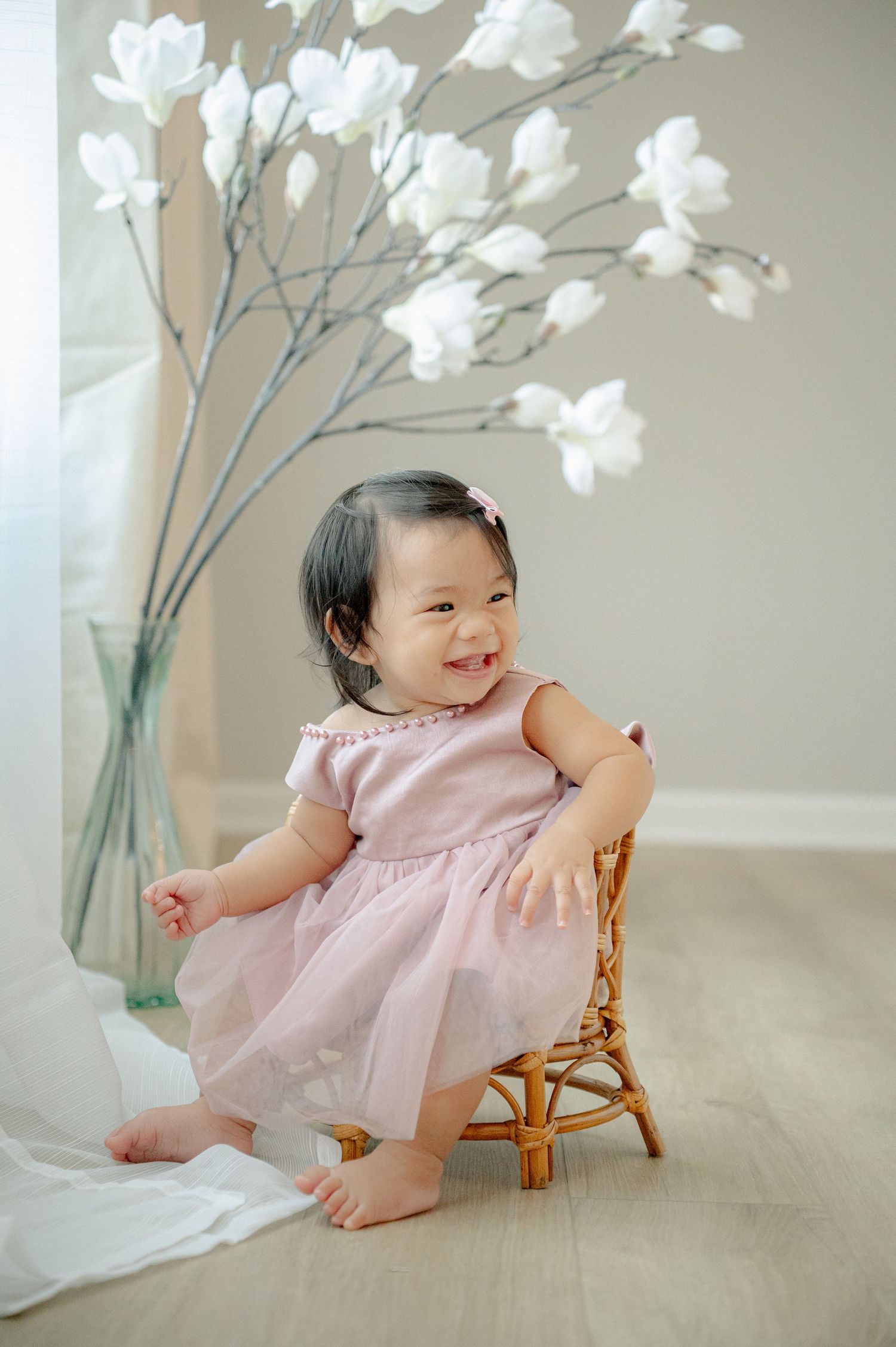 Baby in pink dress sits on rattan chair next to white floral arrangement.