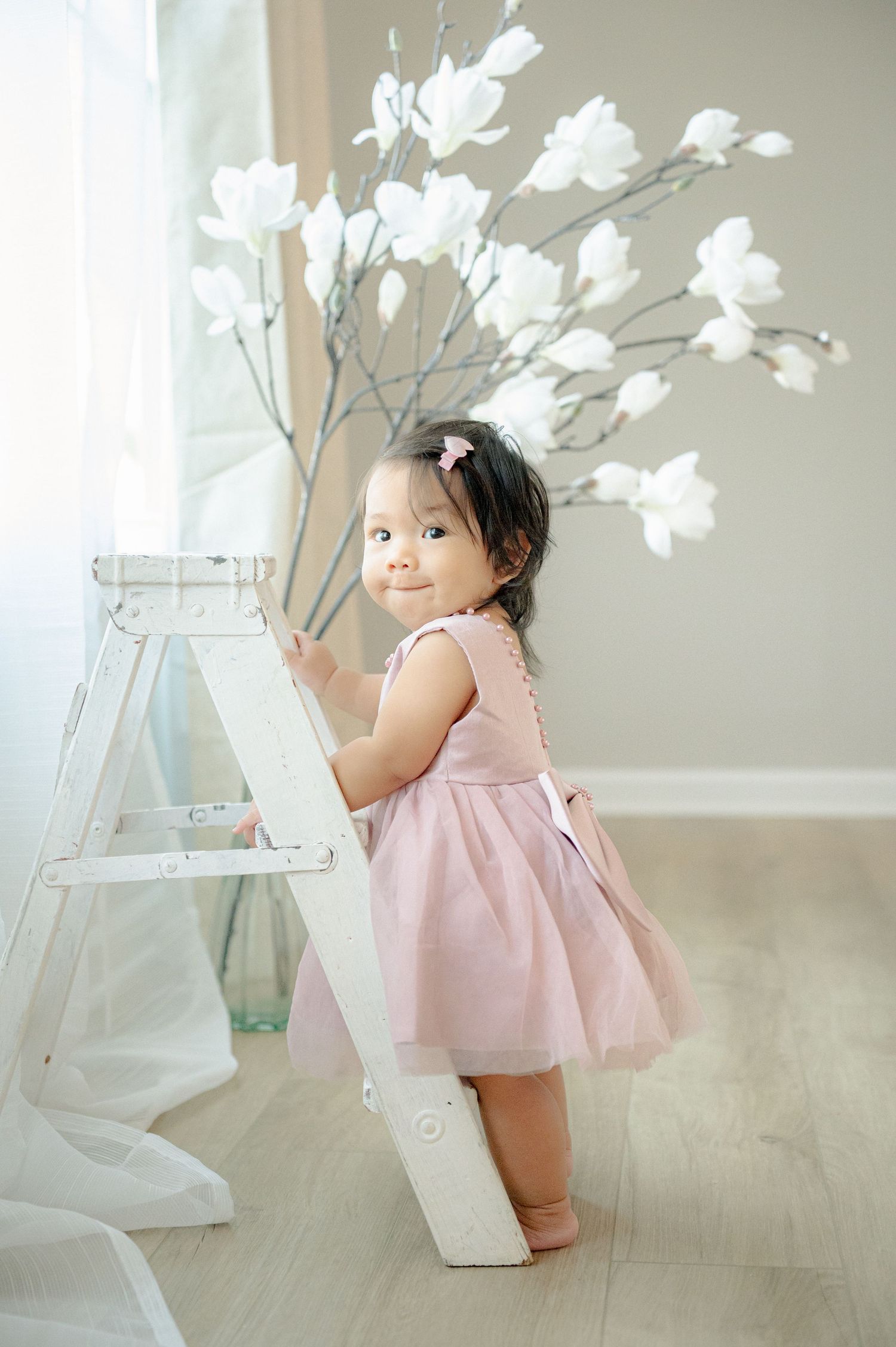 Toddler in pink dress stands near white ladder with decorative flowers.
