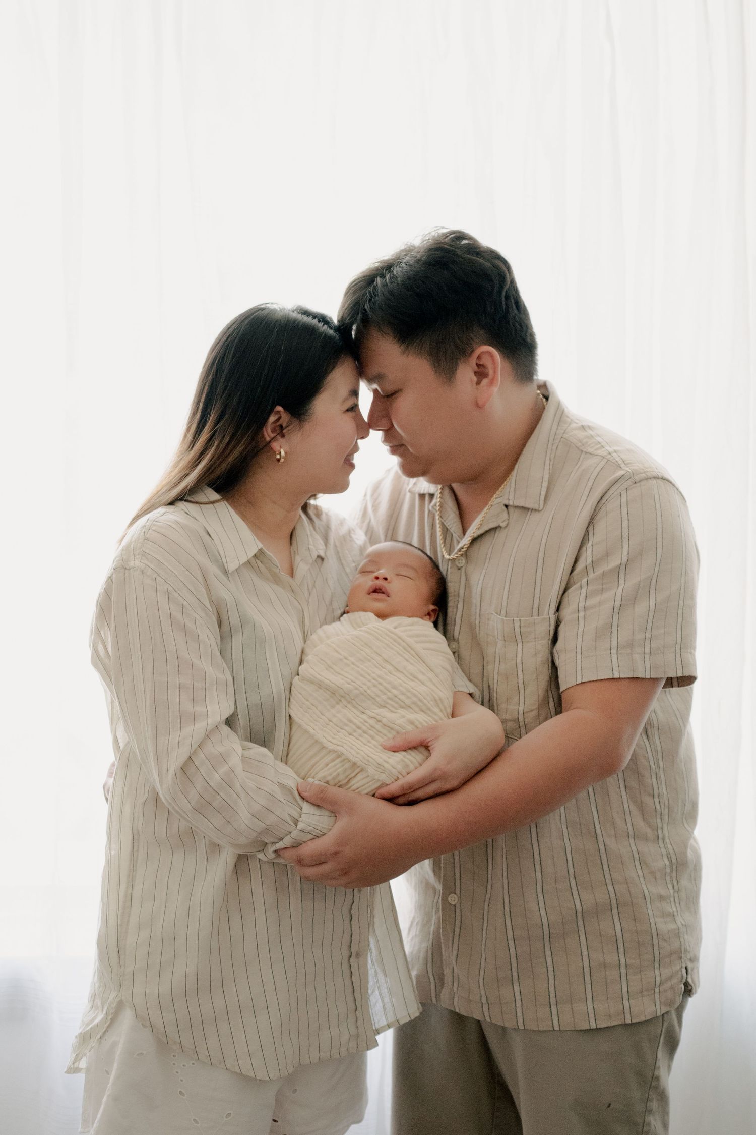 Parents in matching beige outfits share an intimate moment while holding their newborn baby against a white backdrop.