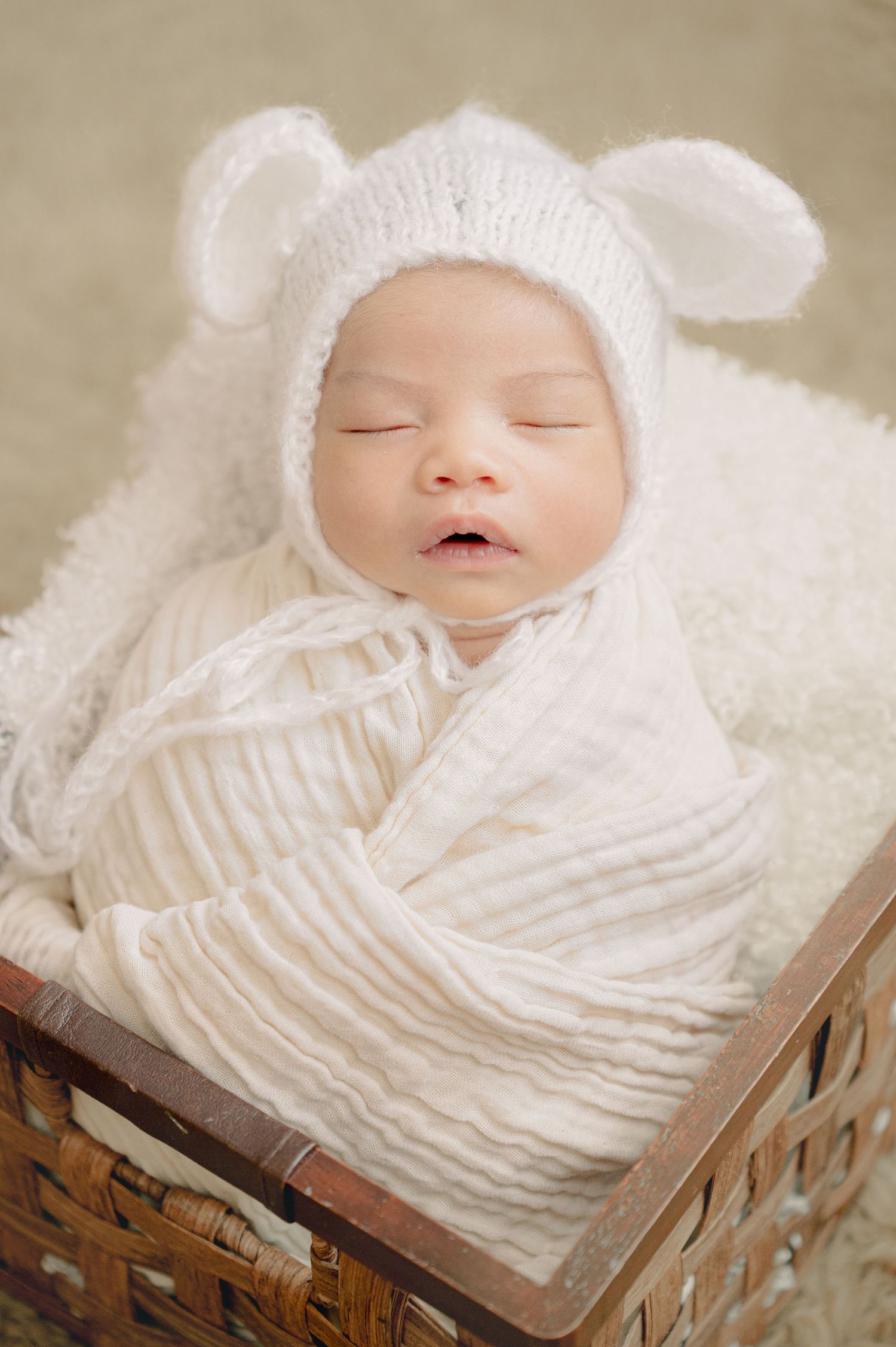Sleeping newborn baby wearing white knit bear hat wrapped in blanket resting in woven basket.