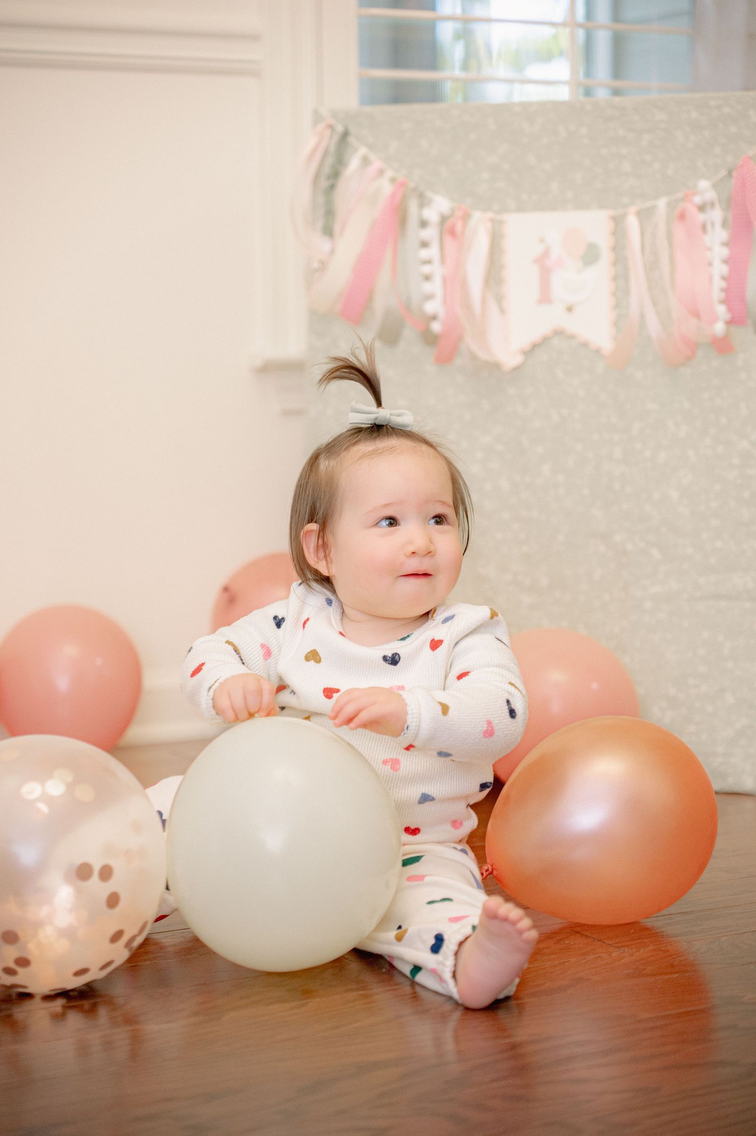 Baby in heart-patterned outfit lying among pink and white balloons on hardwood floor.