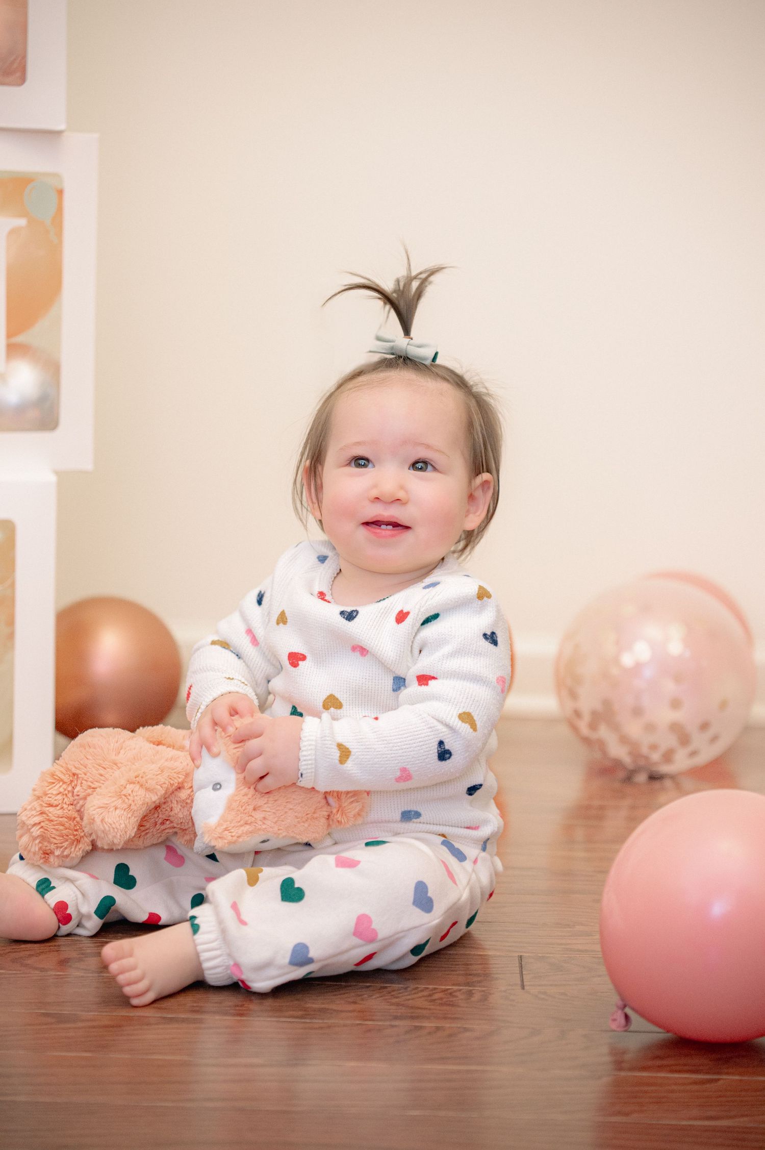 Smiling baby in heart-print pajamas playing with pink and white balloons on wooden floor.