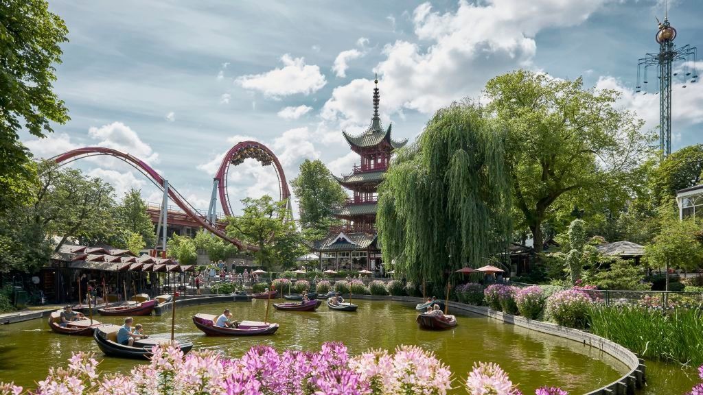 A scenic view of Tivoli Gardens amusement park in Copenhagen with roller coaster tracks, pagoda and blooming flowers by the lake.