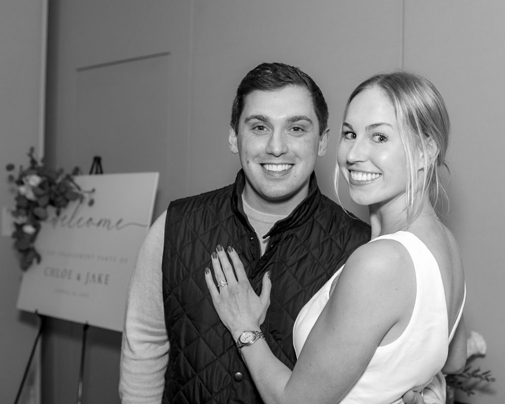 A joyful couple posing at their engagement party next to a decorative welcome sign during engagement party in Westborough, MA. Event Photos