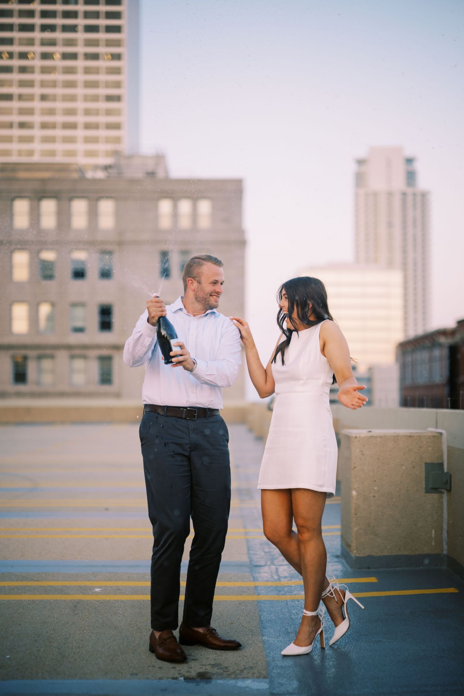 A couple celebrates with champagne on a rooftop parking deck at sunset in the city.