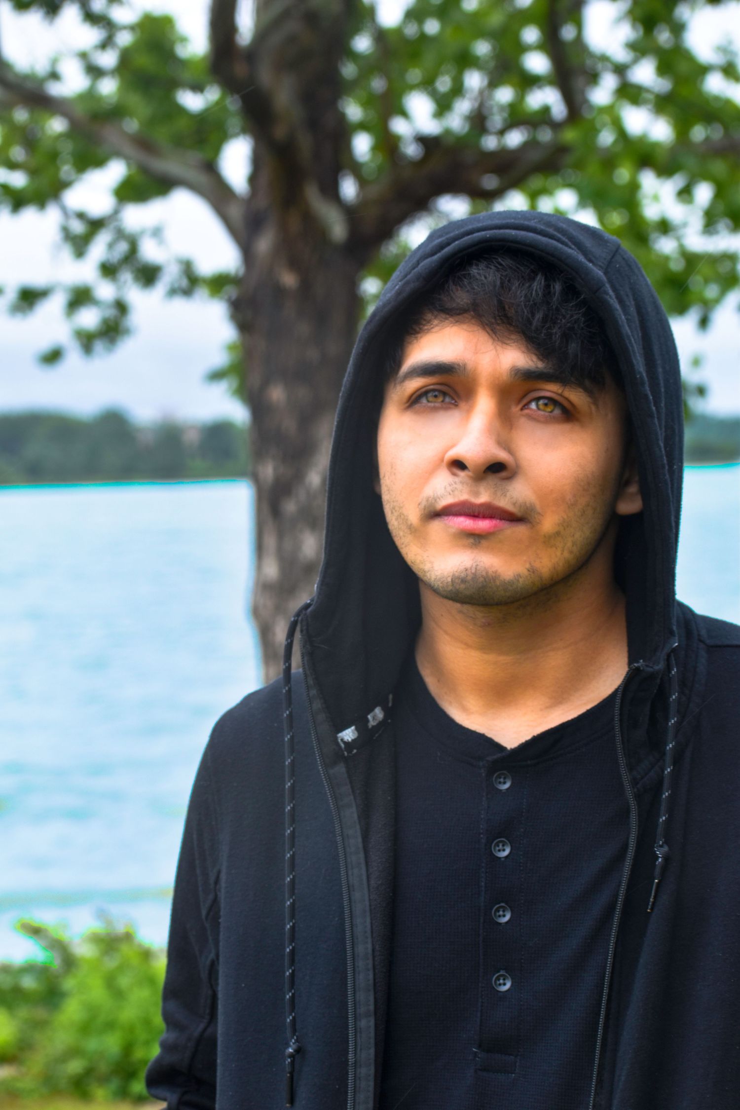 Person wearing a black hooded sweatshirt stands near a lake with trees in the background on an overcast day.