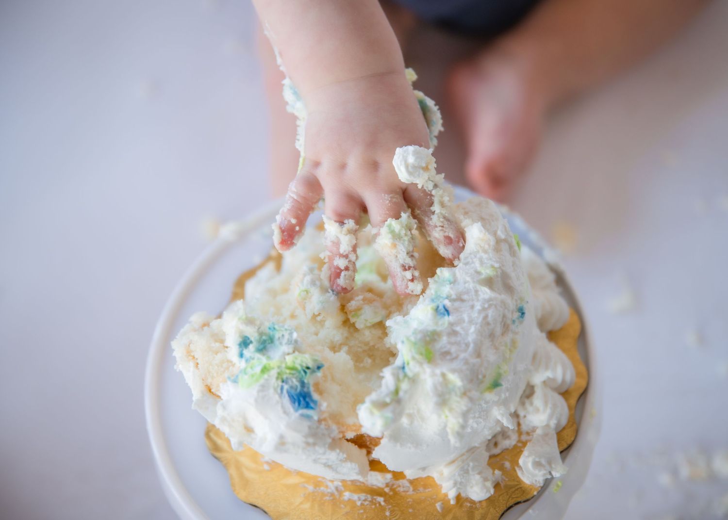 A small hand reaches for a vanilla cake covered in whipped cream and colorful sprinkles.
