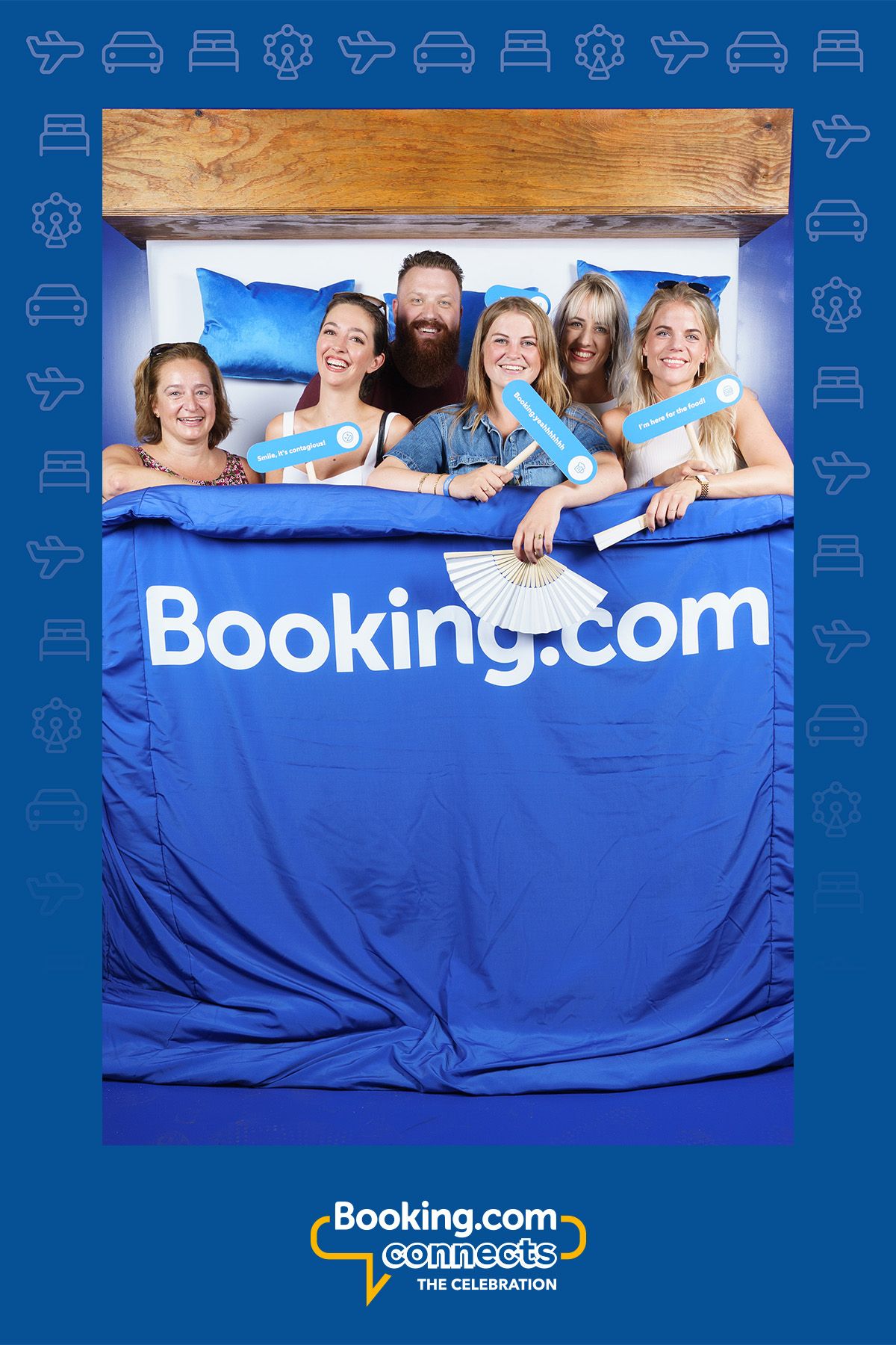 Five smiling people posing in a Booking.com themed photo booth setup with branded blue bedding.