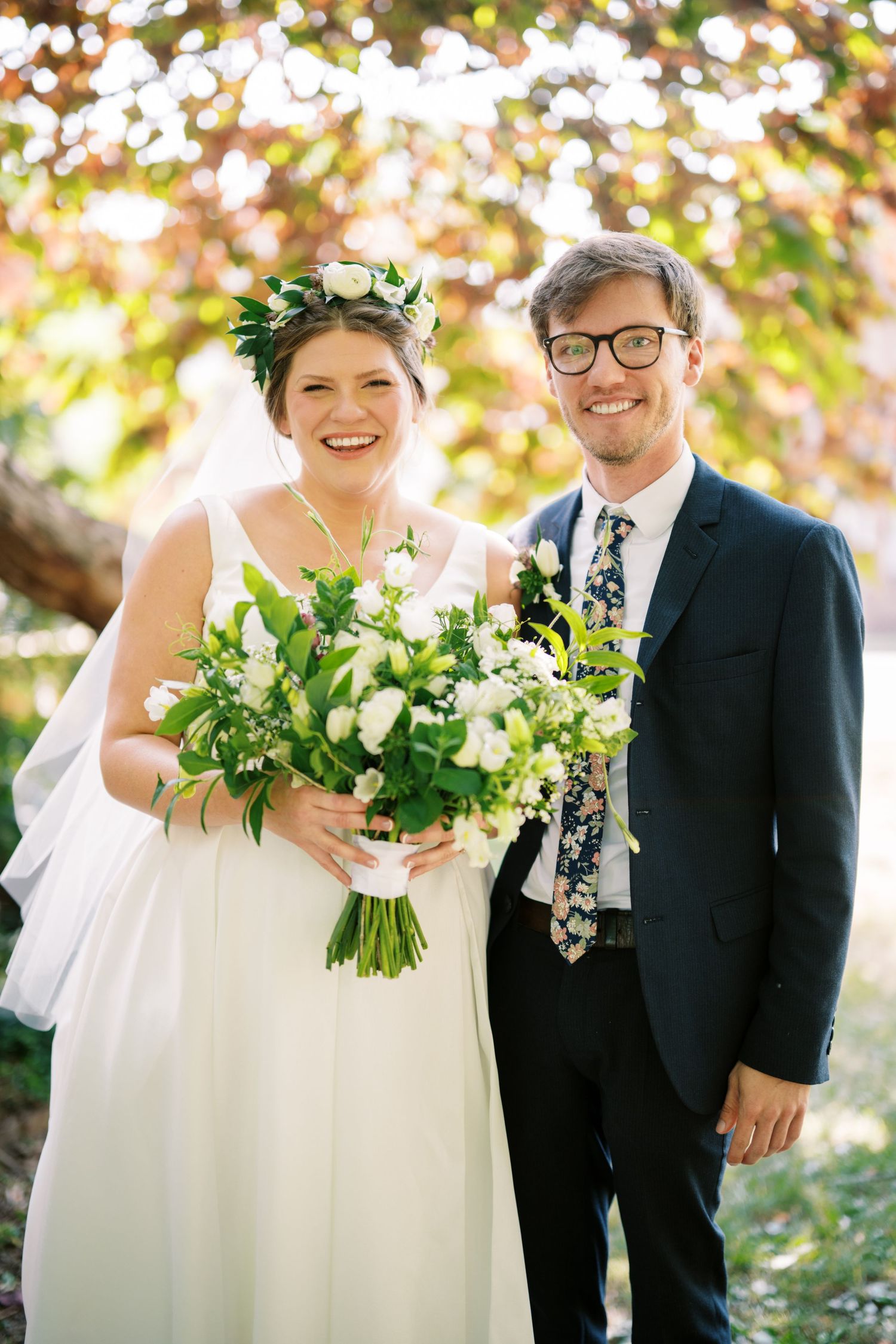 Newlyweds celebrate outdoors with bride in leafy crown and flowing white gown holding lush green and white bouquet.