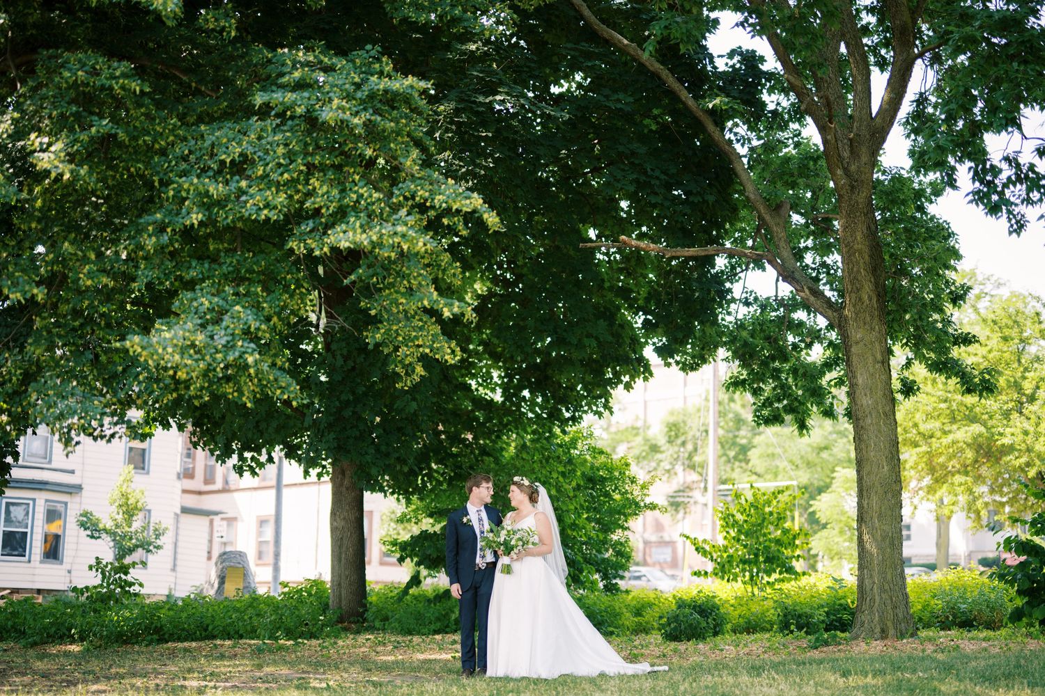 Couple standing together beneath tall green trees in a garden setting on a bright summer day.
