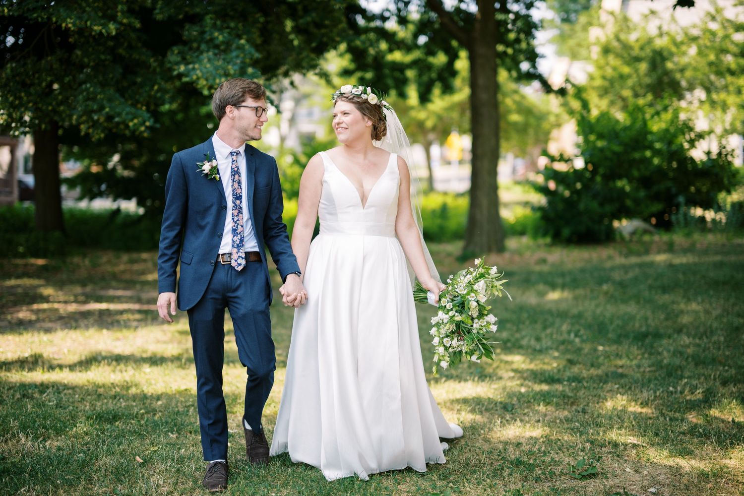 A couple walks hand in hand through a sunlit park on their wedding day, wearing formal attire in a natural setting.