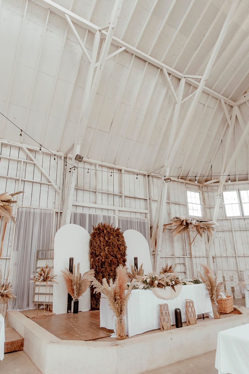 Modern boho wedding reception setup with white archways and dried pampas grass decorations in a white greenhouse venue.
