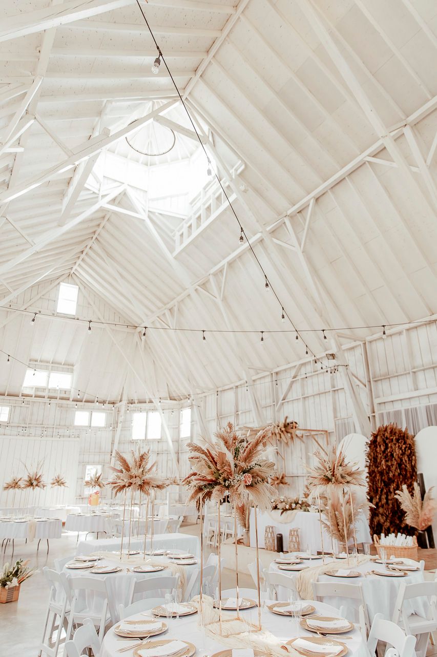 Light-filled white barn venue with suspended pampas grass arrangements and round dining tables set for an event.