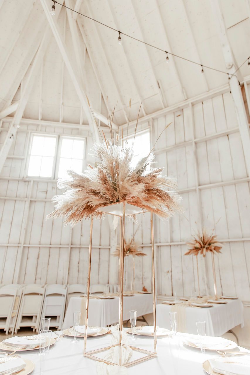 Modern wedding decor featuring tall pampas grass arrangements on gold stands in a white barn-style venue.