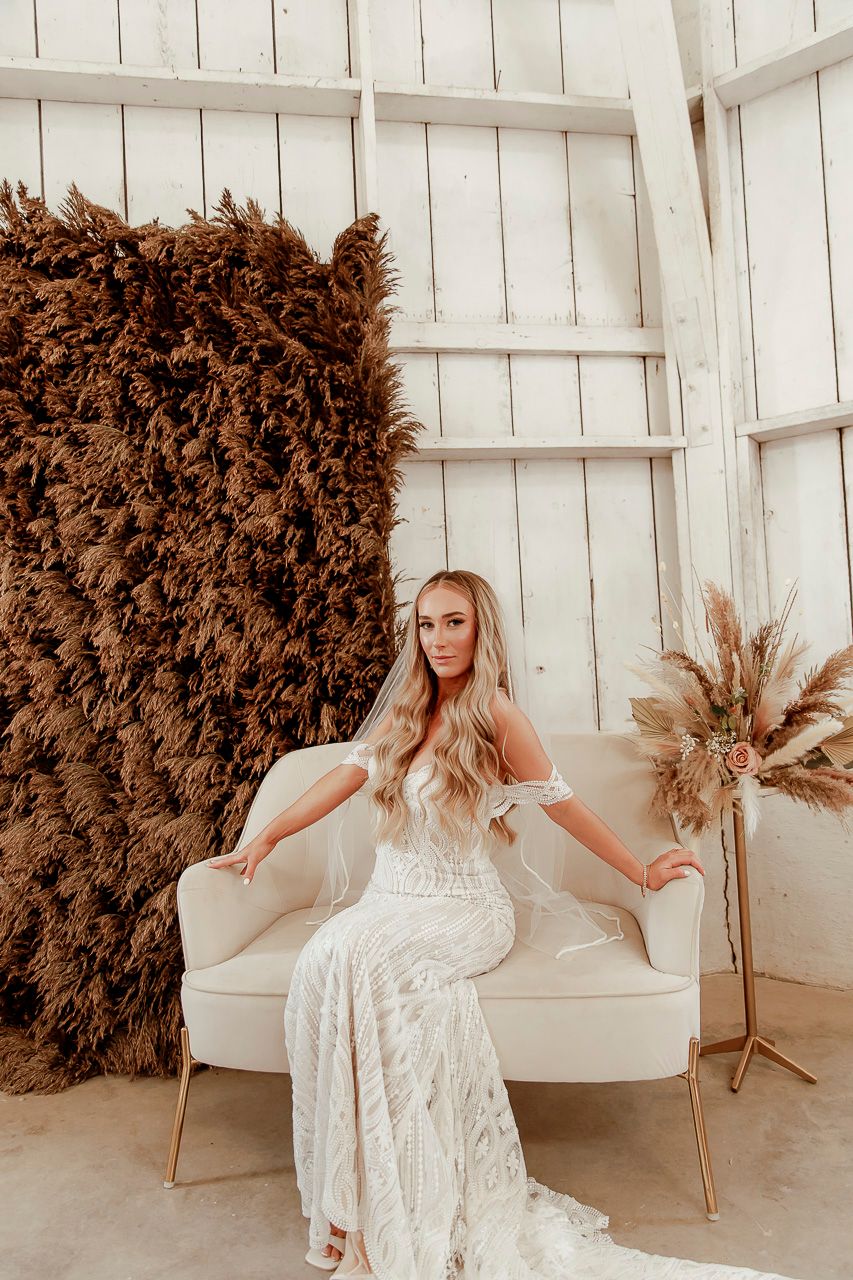 Beautiful lace wedding gown on display in white room with pampas grass and brown textured wall decor.