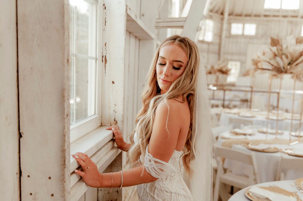 An elegant figure in a white lace wedding gown poses near a sunlit window in a bright, rustic-style room.